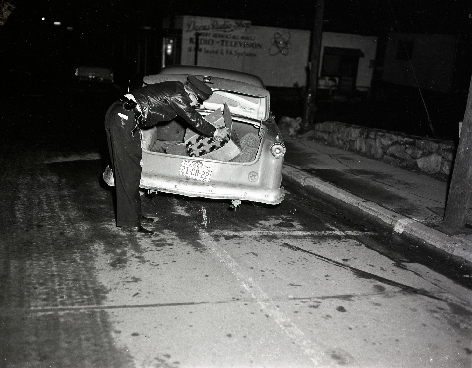 A police officer on the streets of Boone searches a box of jars with clear liquid found in the trunk of a car licensed in Tennessee (date unknown, but probably early 1957). The Dacus Radio Shop is visible in the background. Image is from the forthcoming Palmer Blair Collection, Digital Watauga Project.