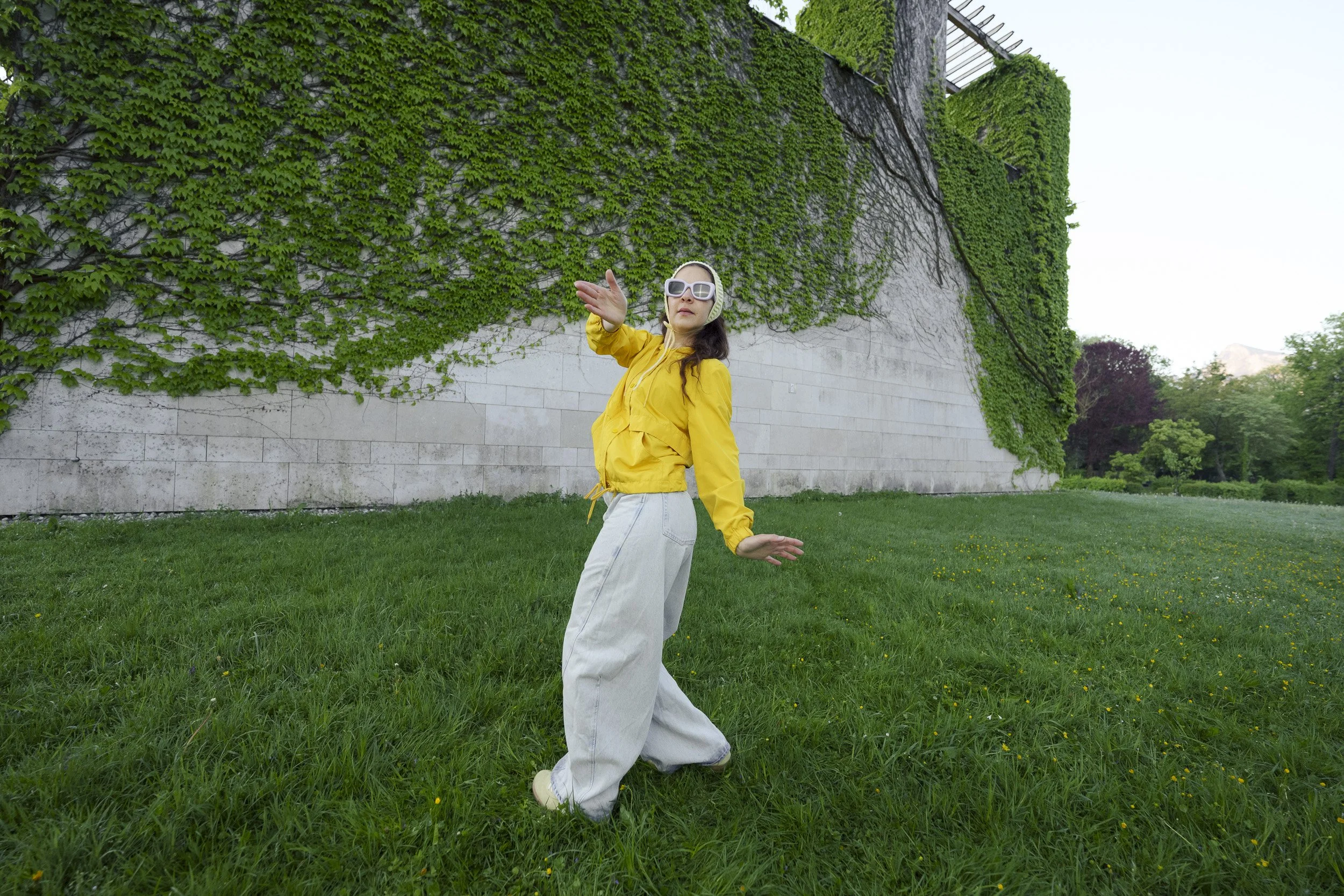 Woman wearing a yellow jacket and white wide-leg pants posing outdoors in front of a stone wall covered in green ivy, with trees in the background.