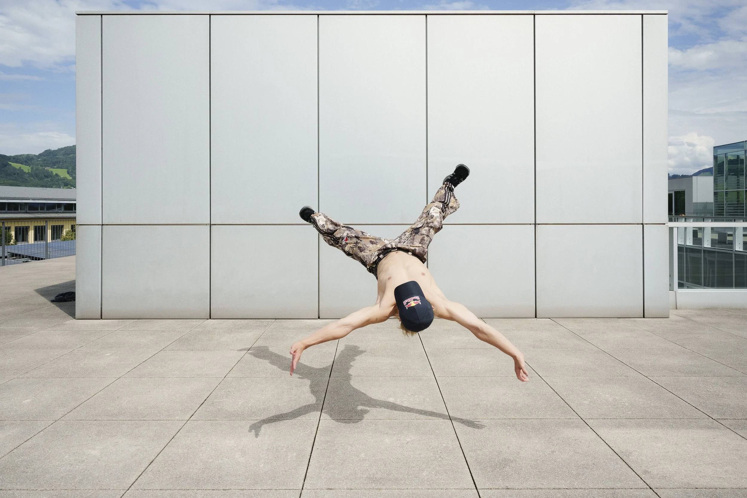 Person performing a handstand on an outdoor concrete surface with a modern building and cloudy sky in the background.
