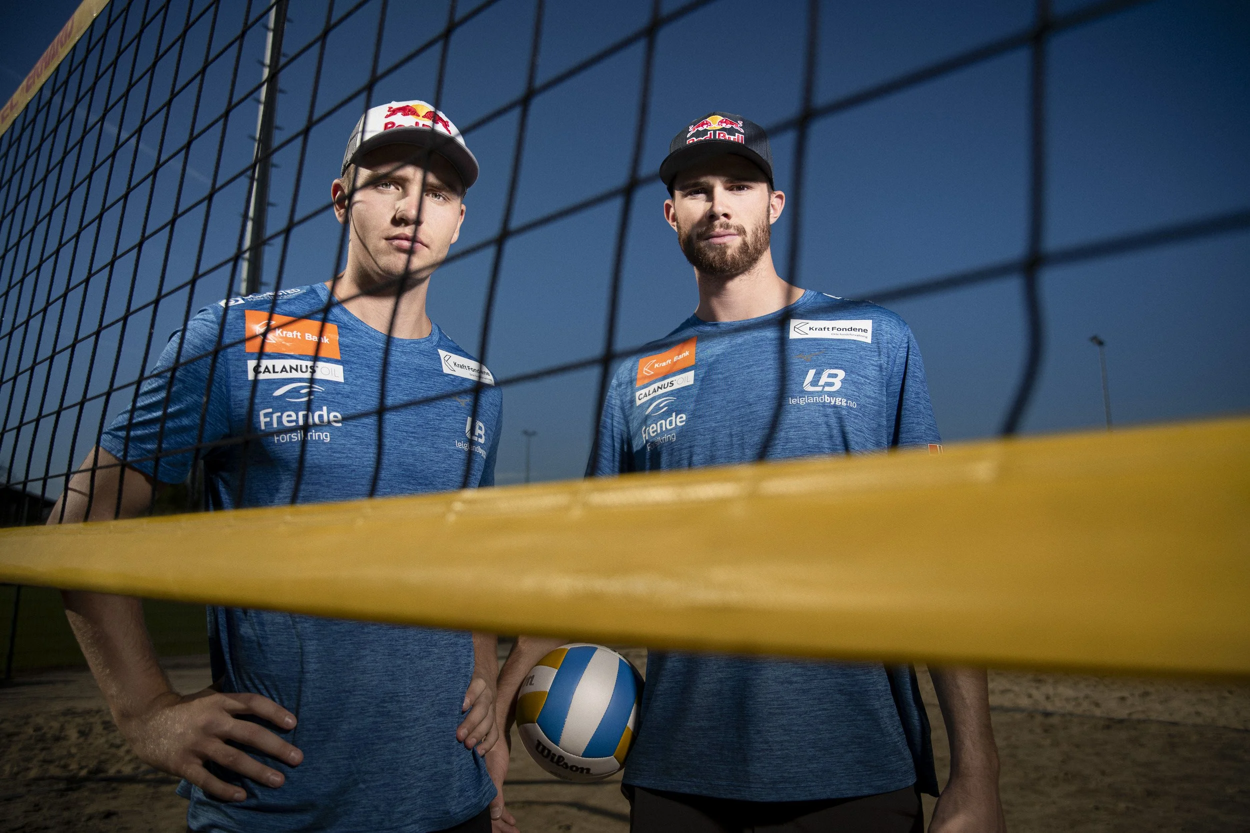 Two male beach volleyball players standing behind a yellow volleyball net, posing for a photo on a sand court at dusk. They are wearing blue sports shirts with various sponsor logos and black caps.