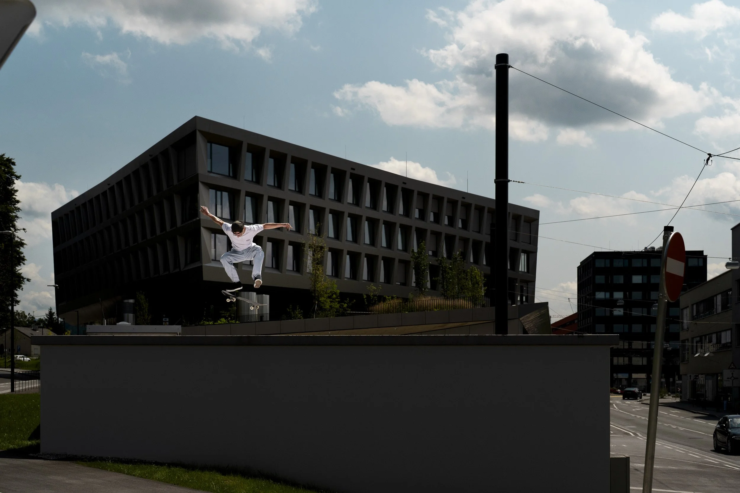 A skateboarder performing a trick in mid-air against an urban background with modern buildings, a cloudy sky, utility poles, and a street with cars.