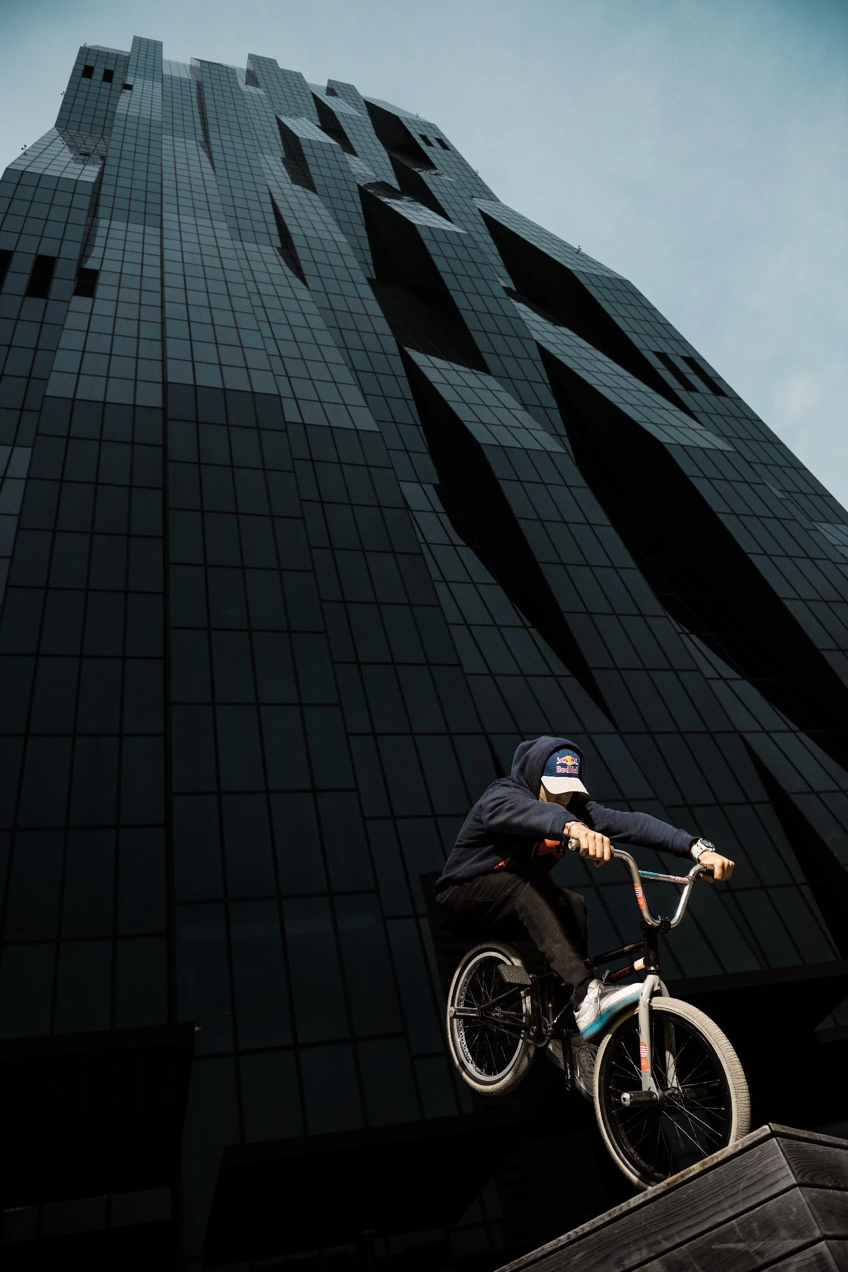 A person wearing a hoodie and cap riding a BMX bike on a ledge against a tall, dark modern glass building.