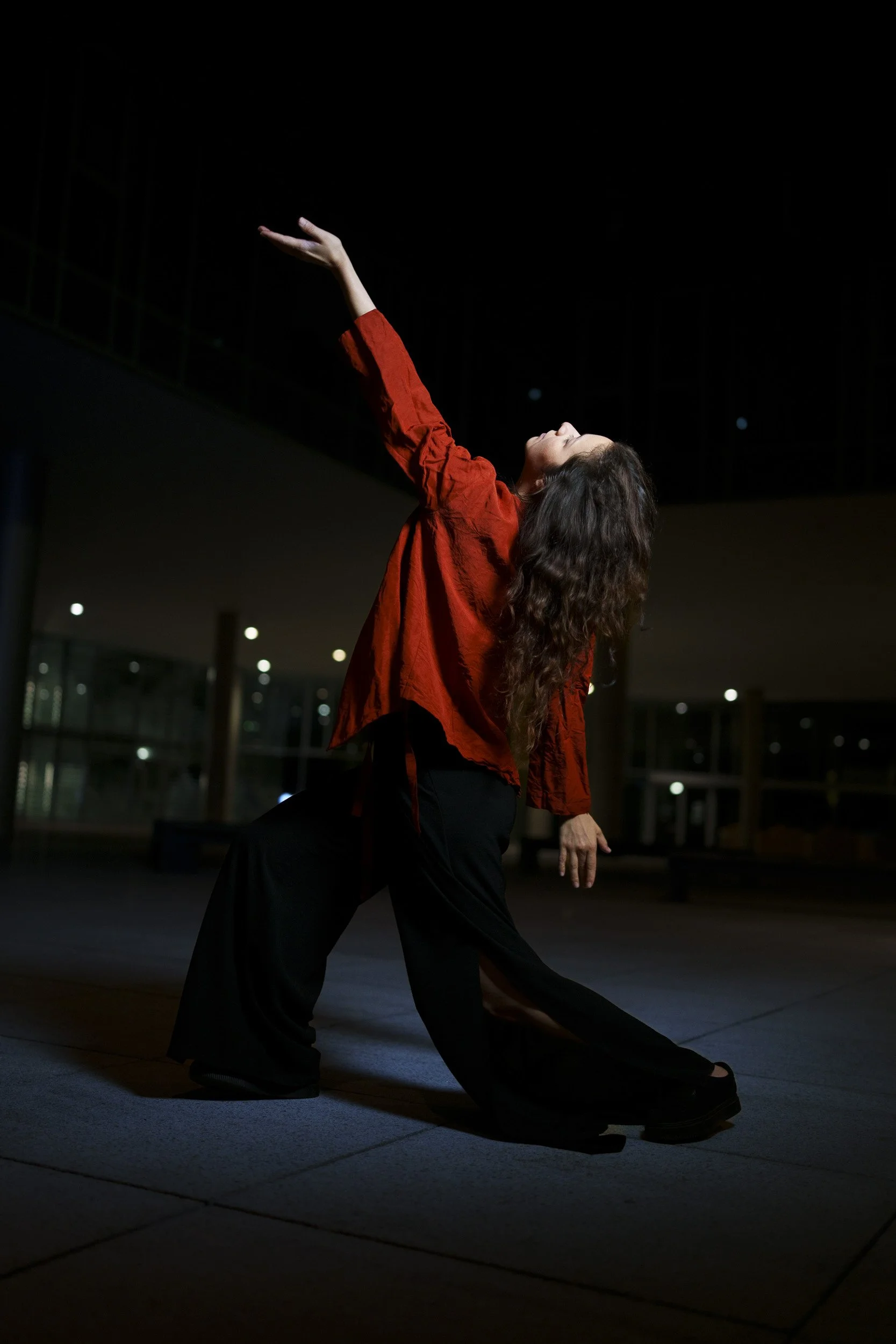 A woman with curly hair wearing a red shirt and black pants poses dramatically at night indoors, extending her right arm upward and leaning back.