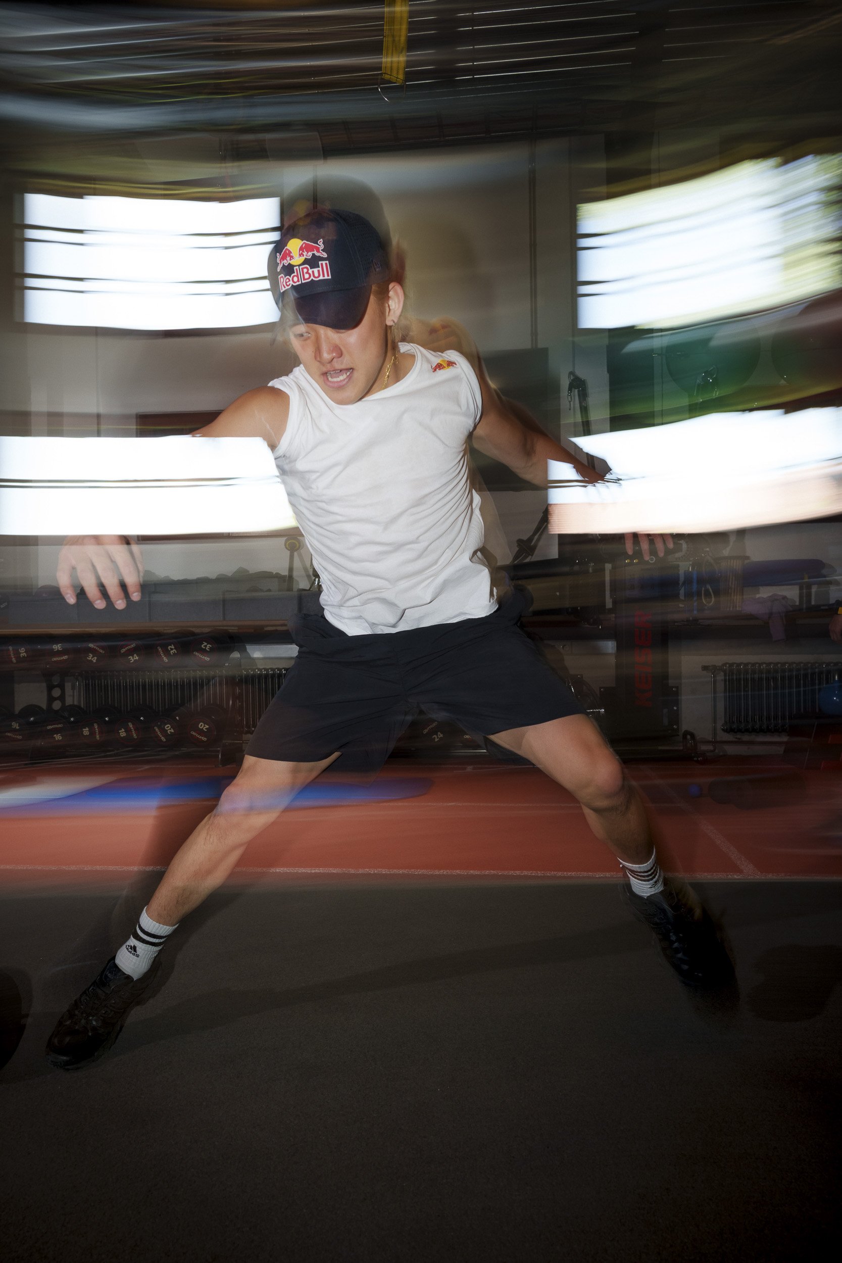 A woman wearing a Red Bull cap and athletic clothes is in motion, lunging with bent knees and outstretched arms inside a gym.