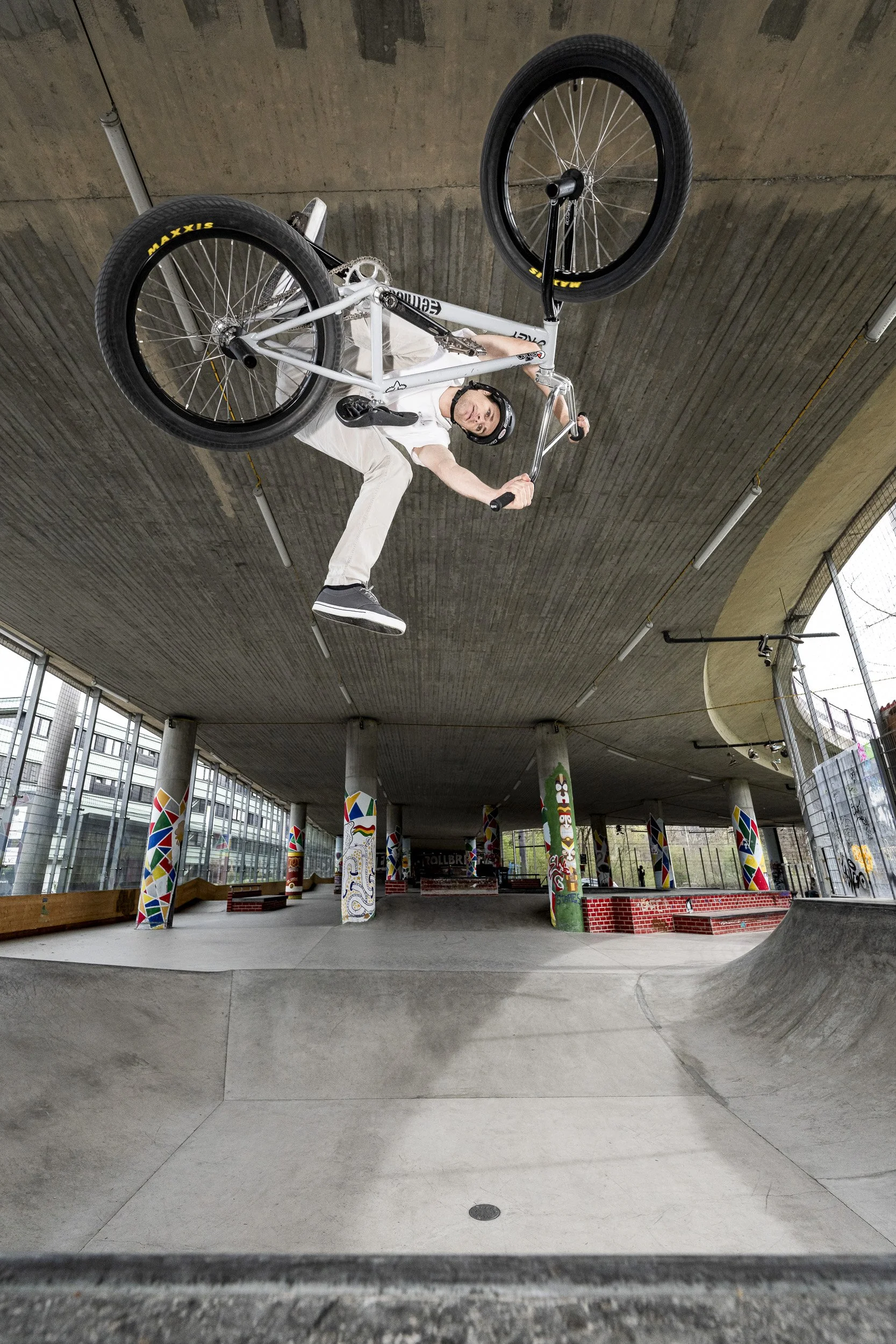 A man performing an aerial BMX stunt upside down in a skate park, wearing a helmet, white t-shirt, and beige pants.