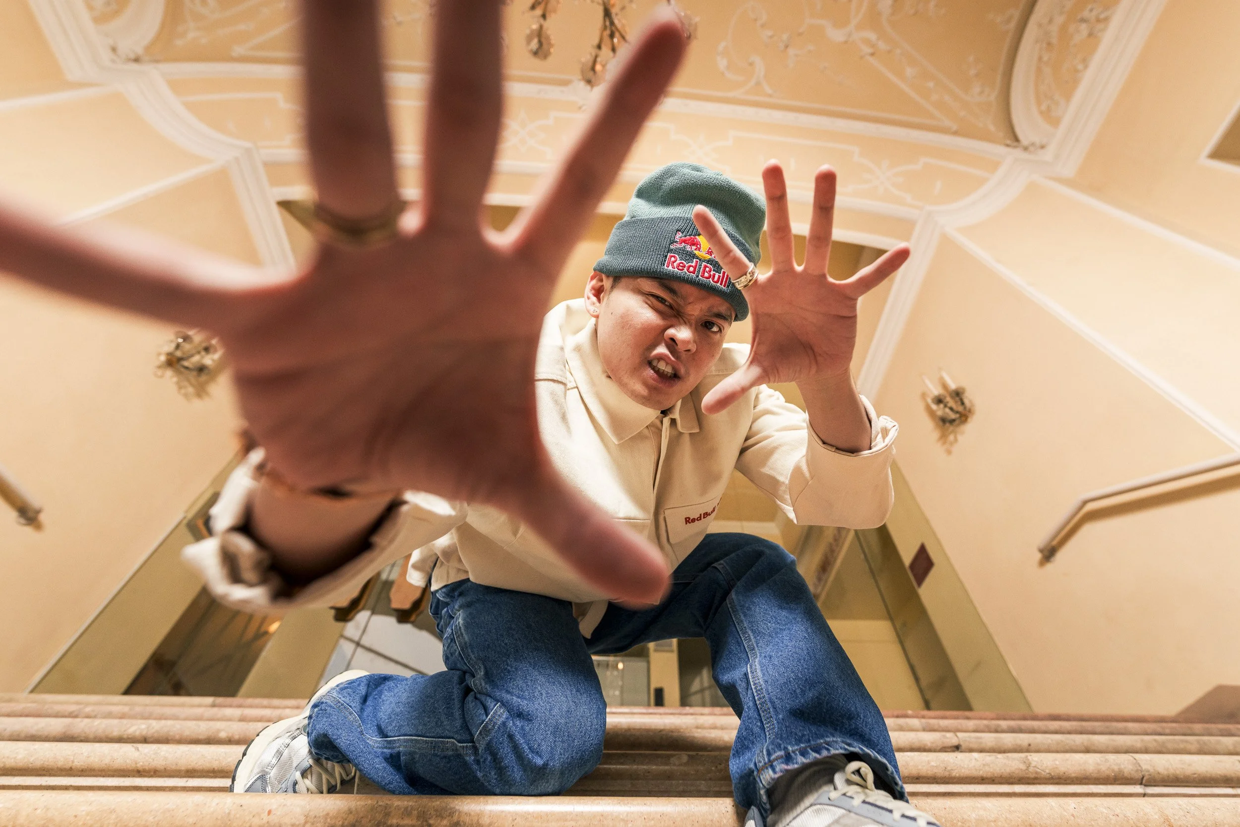 A person crouching on a staircase reaching towards the camera with an intense expression, wearing a green beanie and a beige Red Bull jacket in an ornate room.