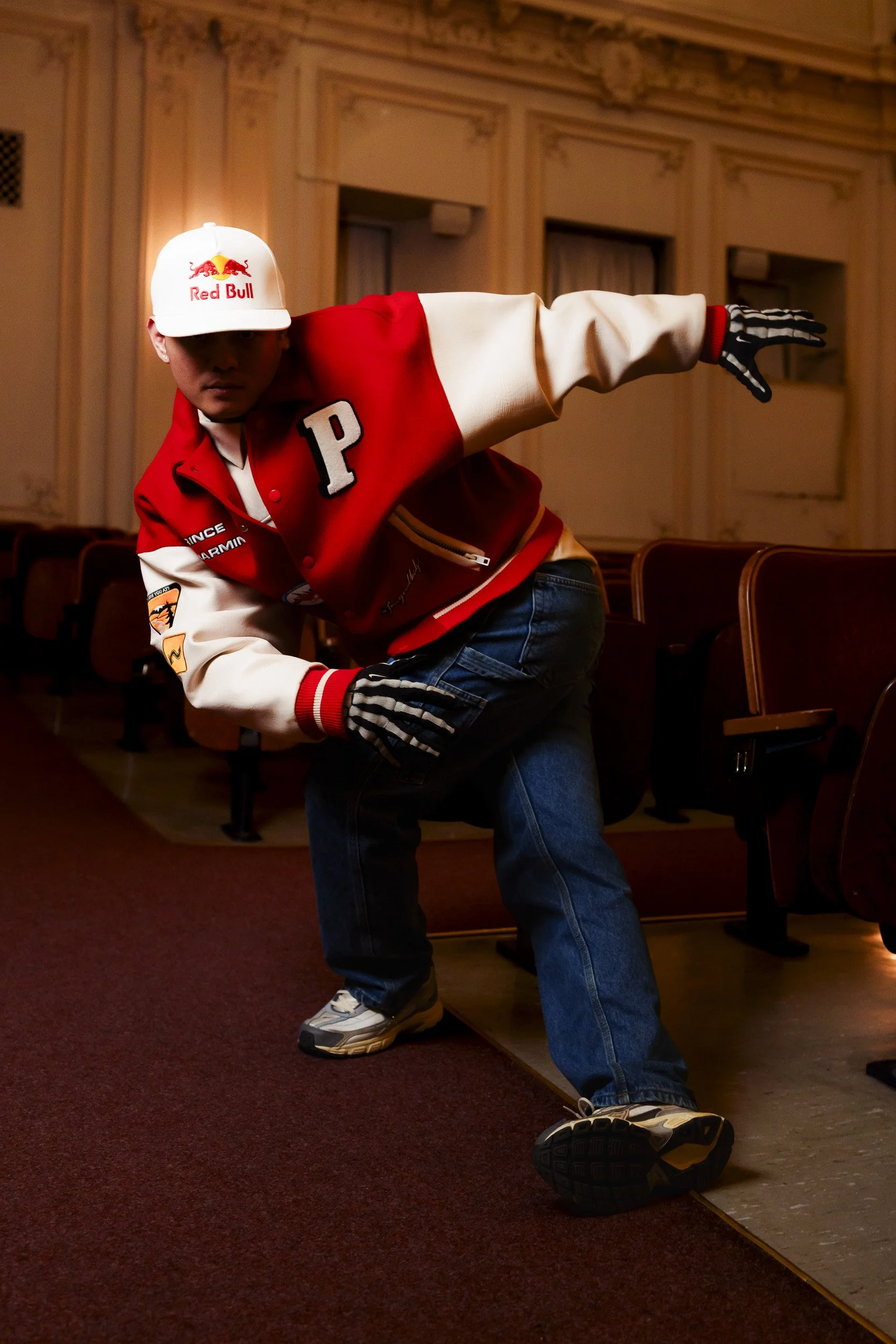 A person in a Red Bull branded white cap and a red and white varsity jacket is striking a dance pose in an ornate room with vintage theater seats.