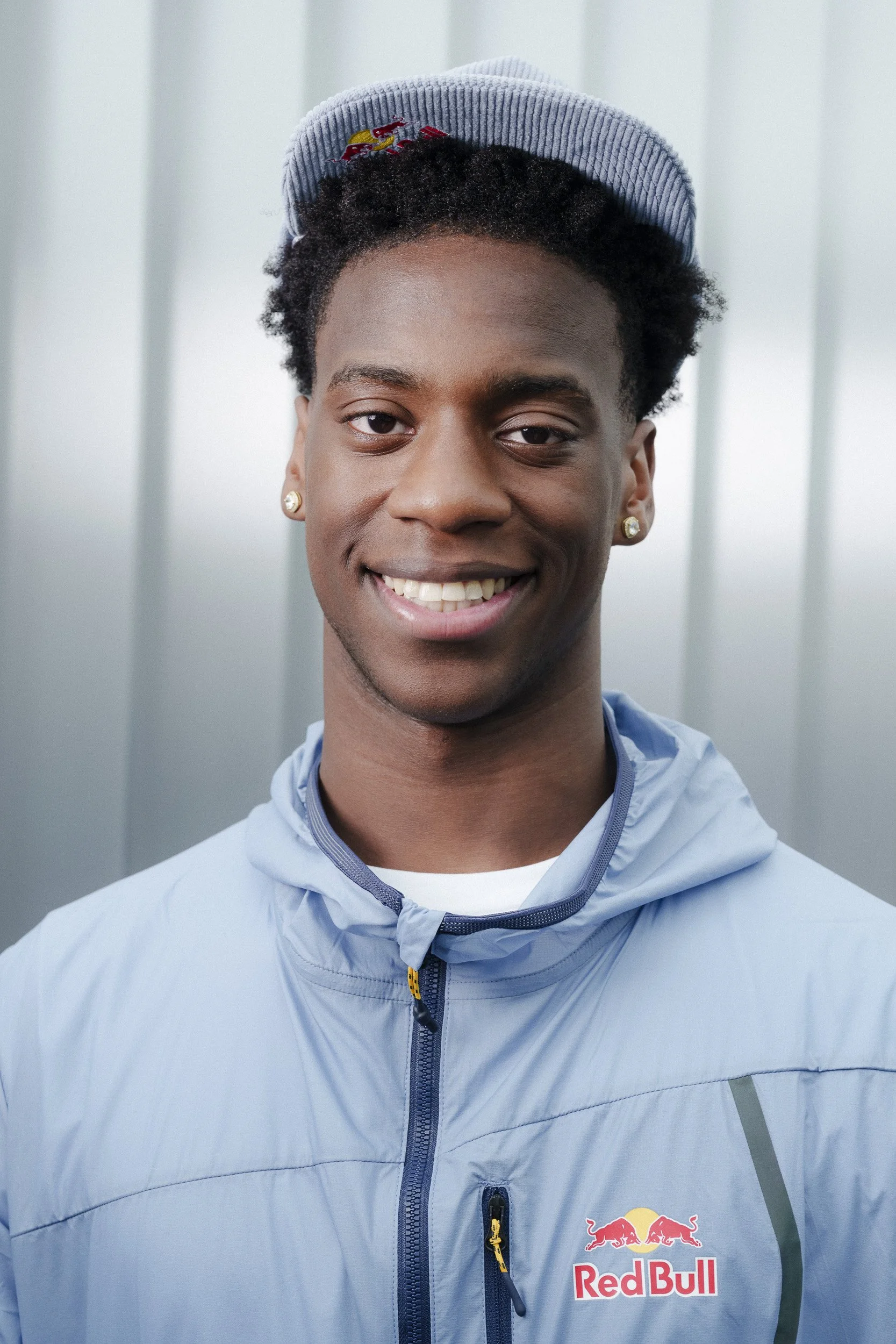 A young man smiling, wearing a light gray jacket with a Red Bull logo and a blue striped cap, standing against a metallic background.