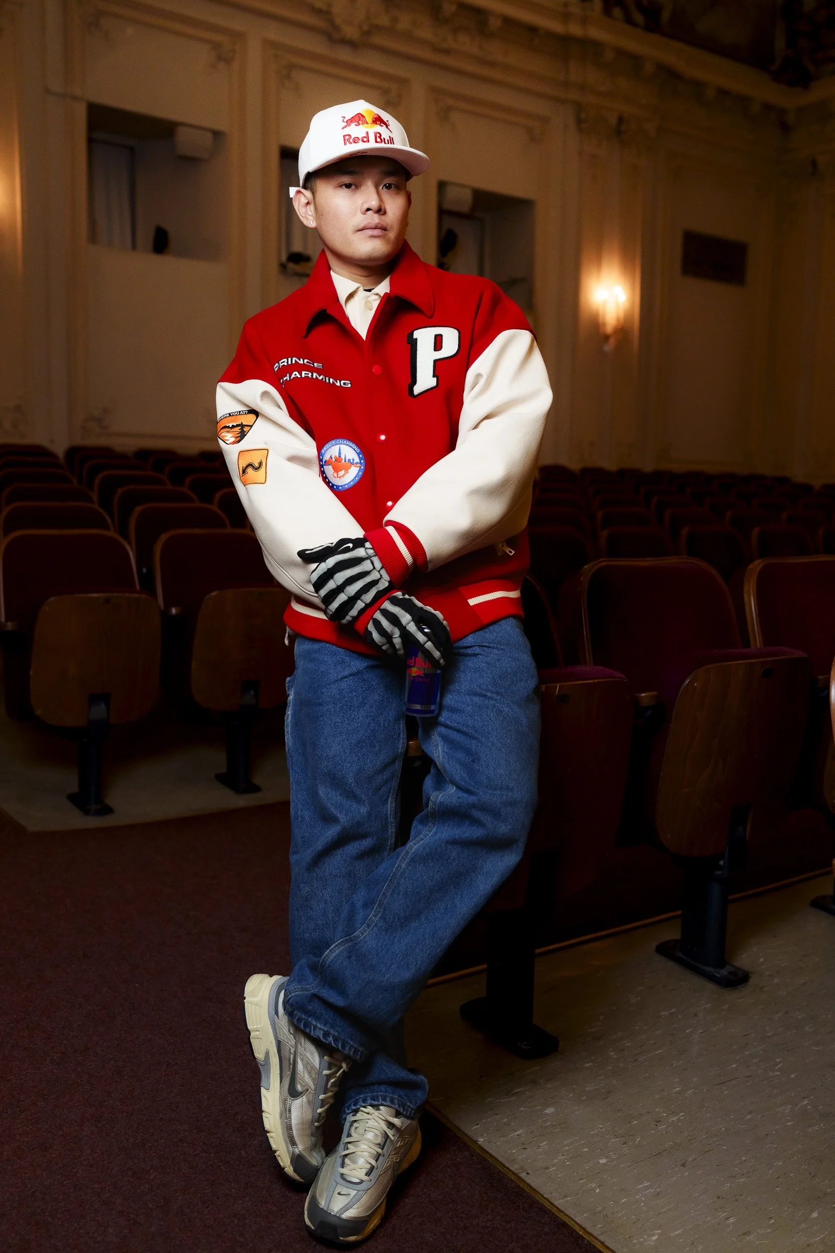 A young man standing in an auditorium, wearing a red and white varsity jacket, blue jeans, sneakers, a white cap with a Red Bull logo, and striped gloves.
