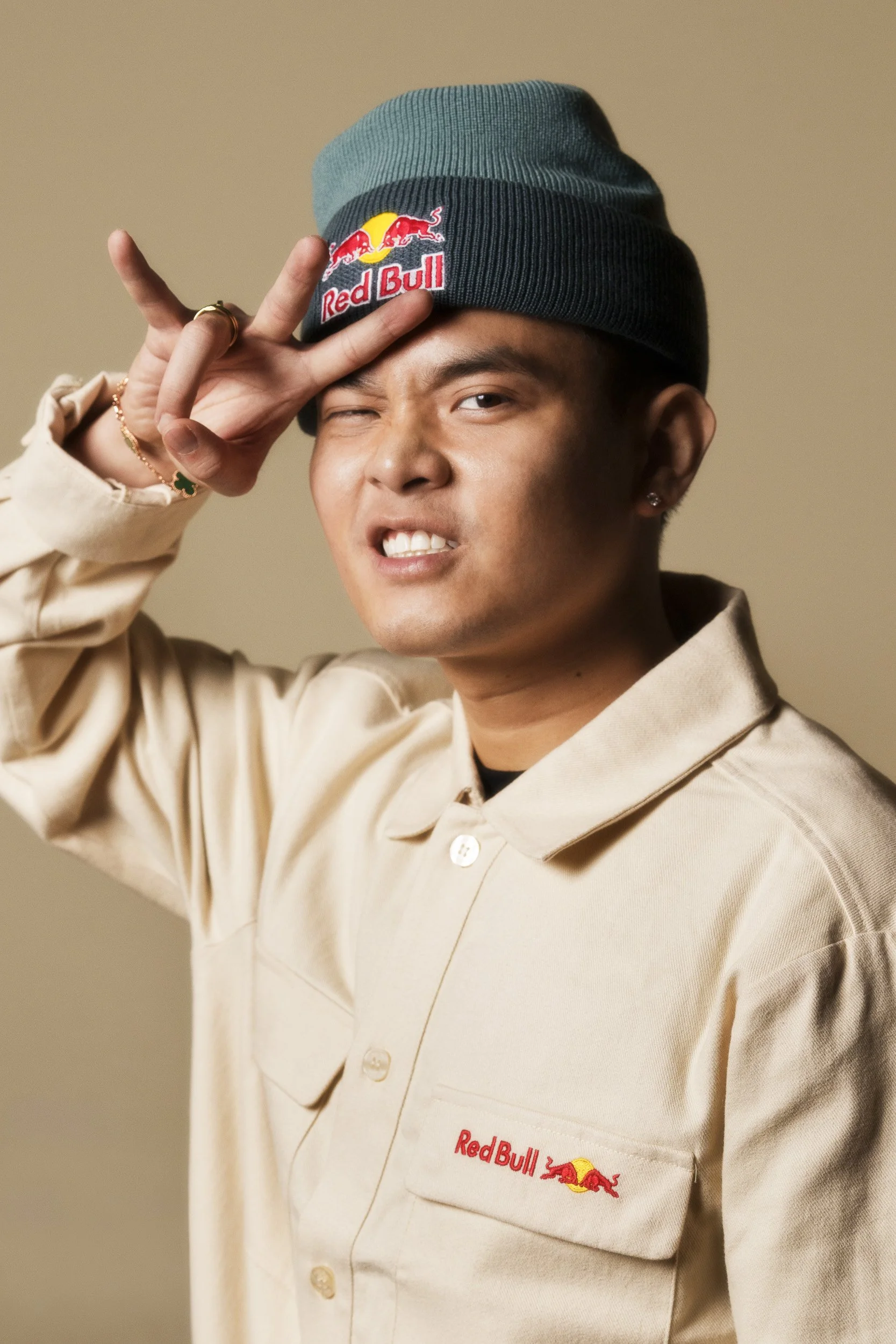 A person is posing with a playful expression, wearing a Red Bull beanie and a beige jacket with the Red Bull logo. They are making a peace sign near their face with one hand.