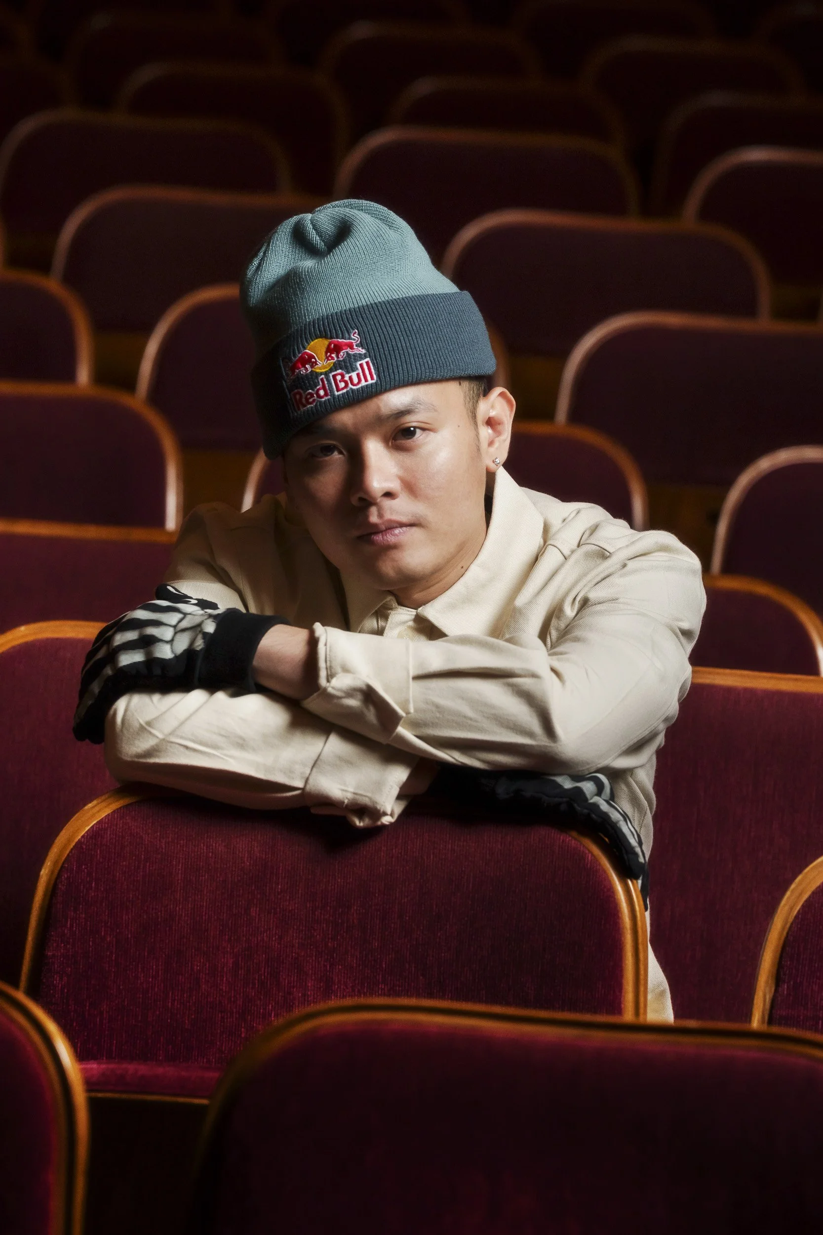 A man wearing a beige jacket and a beanie with the Red Bull logo, sitting in an empty theater with red seats, resting his arms on a seat in front of him.
