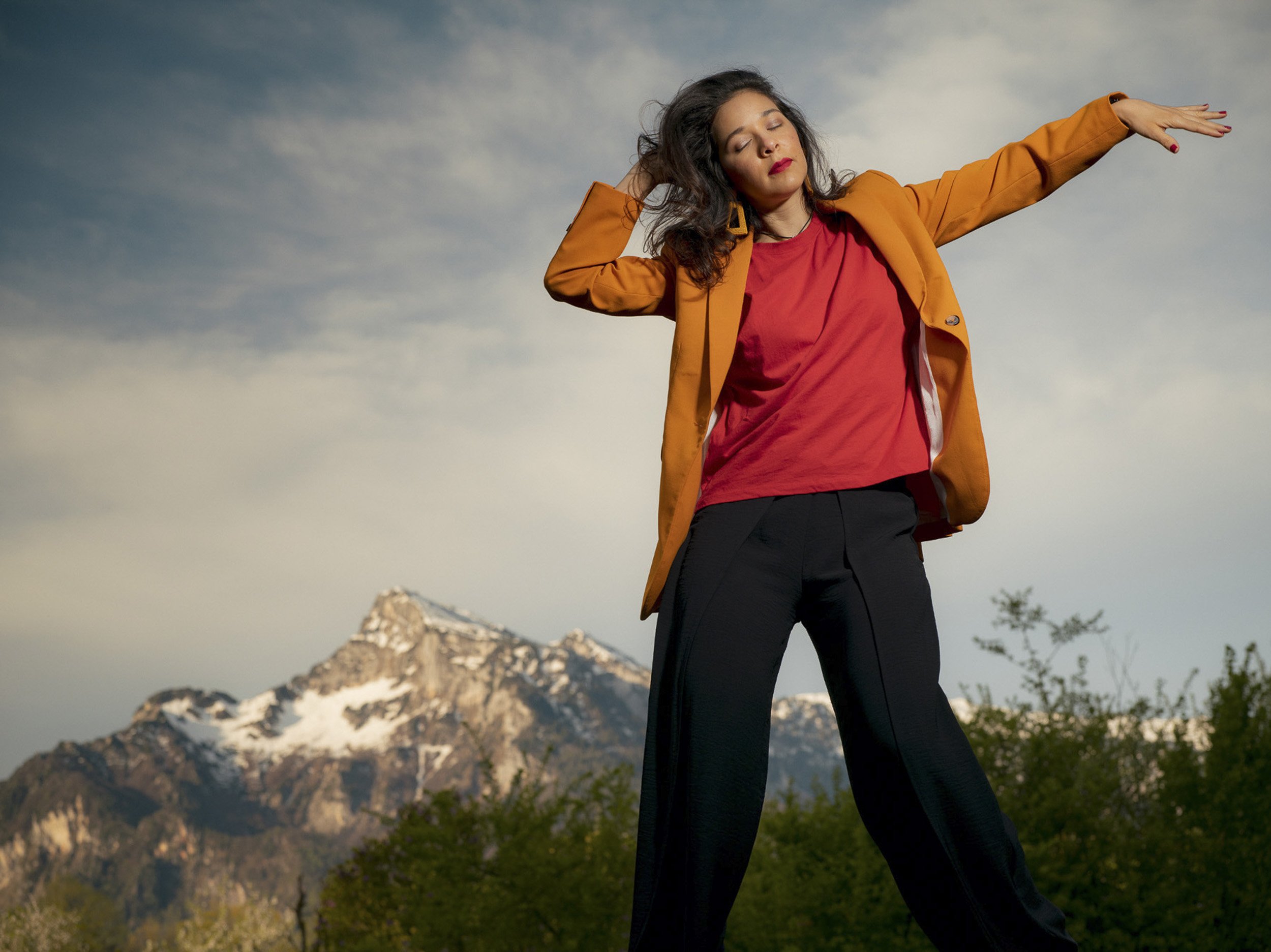 Woman with closed eyes and relaxed expression, standing outdoors with mountain background, wearing an orange jacket, red shirt, and black pants, extending arms to the sides.