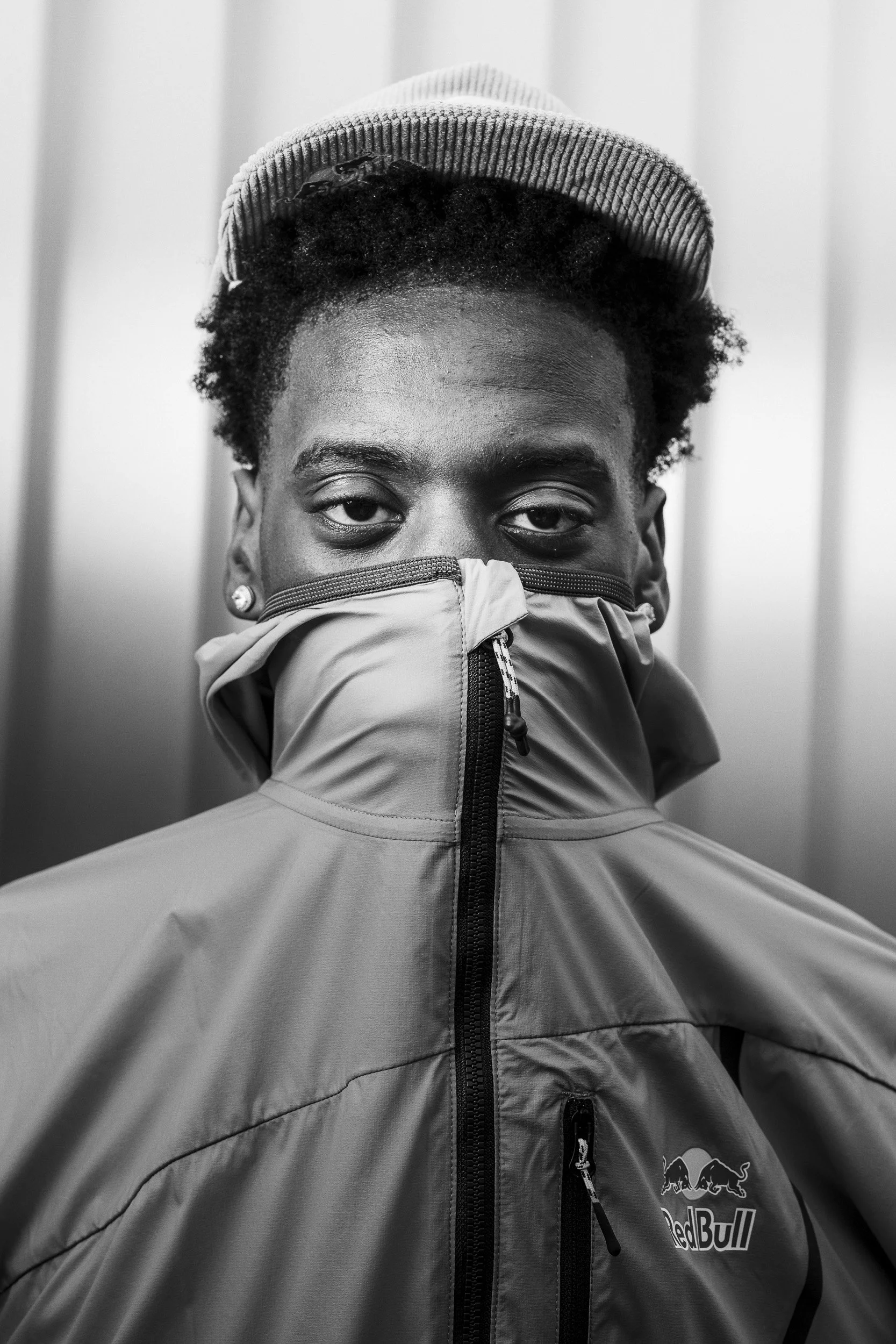 A person wearing a beanie, jacket, and face covering, looking directly at the camera in black and white.