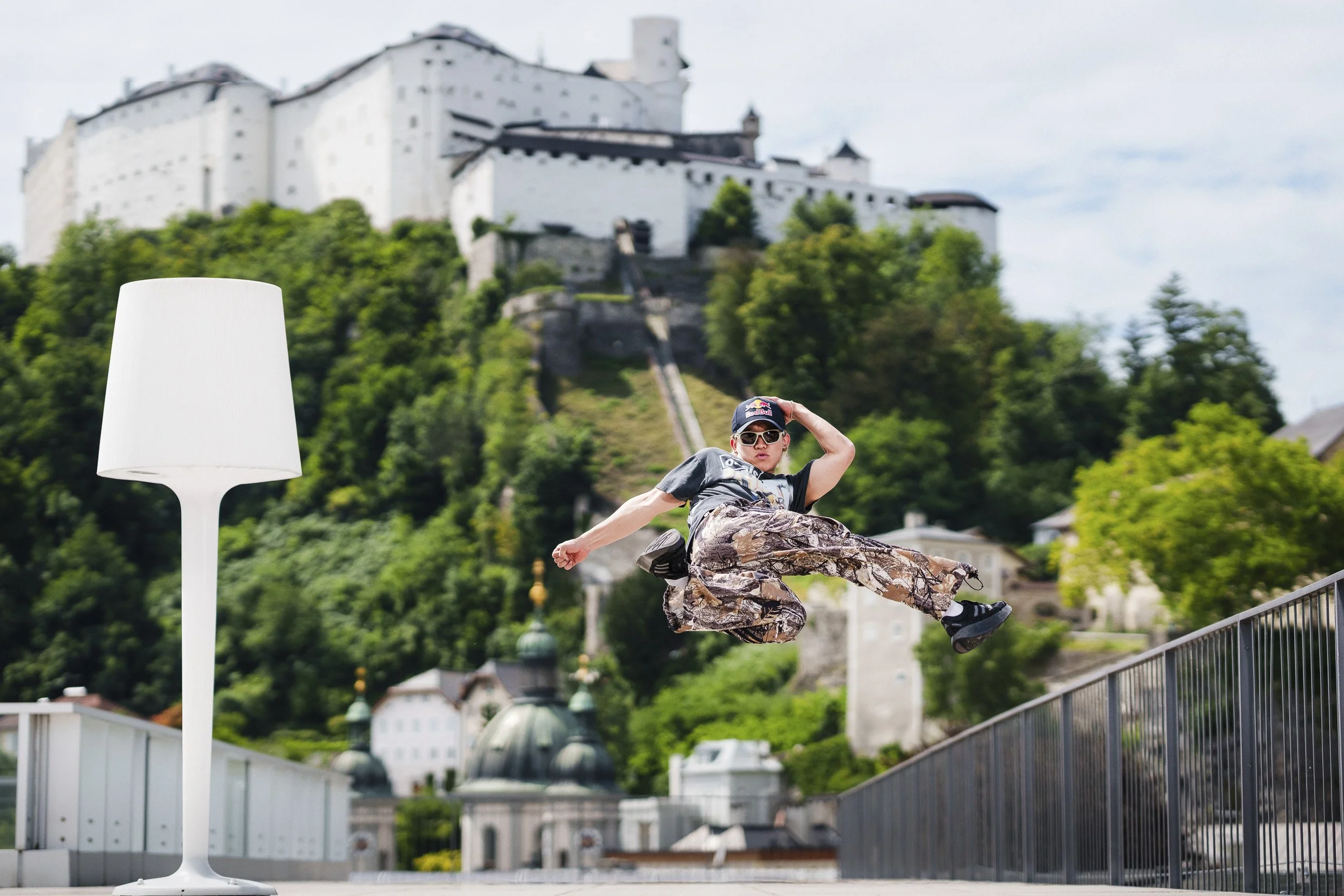 A person mid-air jump next to a modern outdoor lamp post with a castle on a hill in the background.