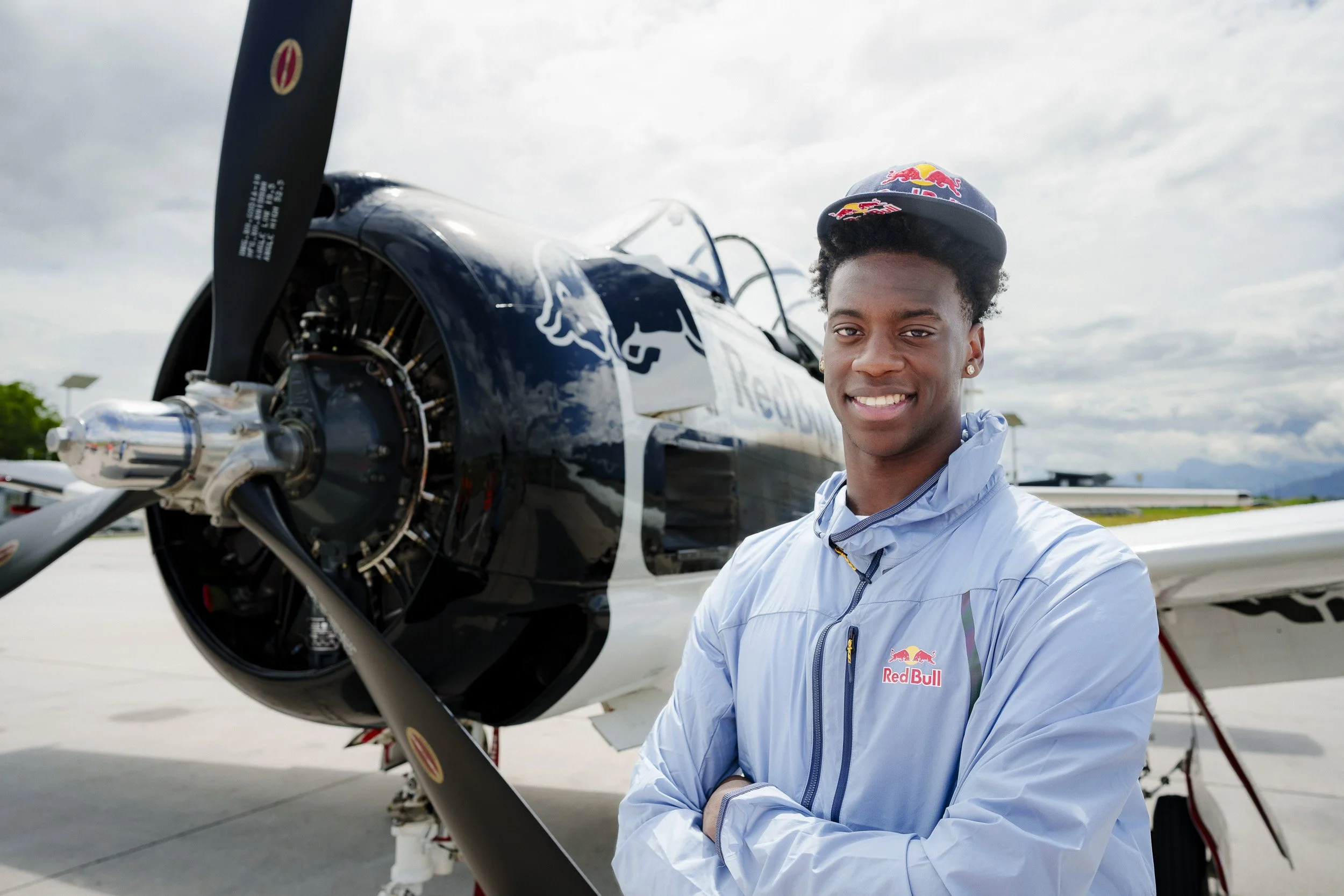 A young man in a Red Bull jacket and cap standing in front of a race plane at an airport, smiling with arms crossed.