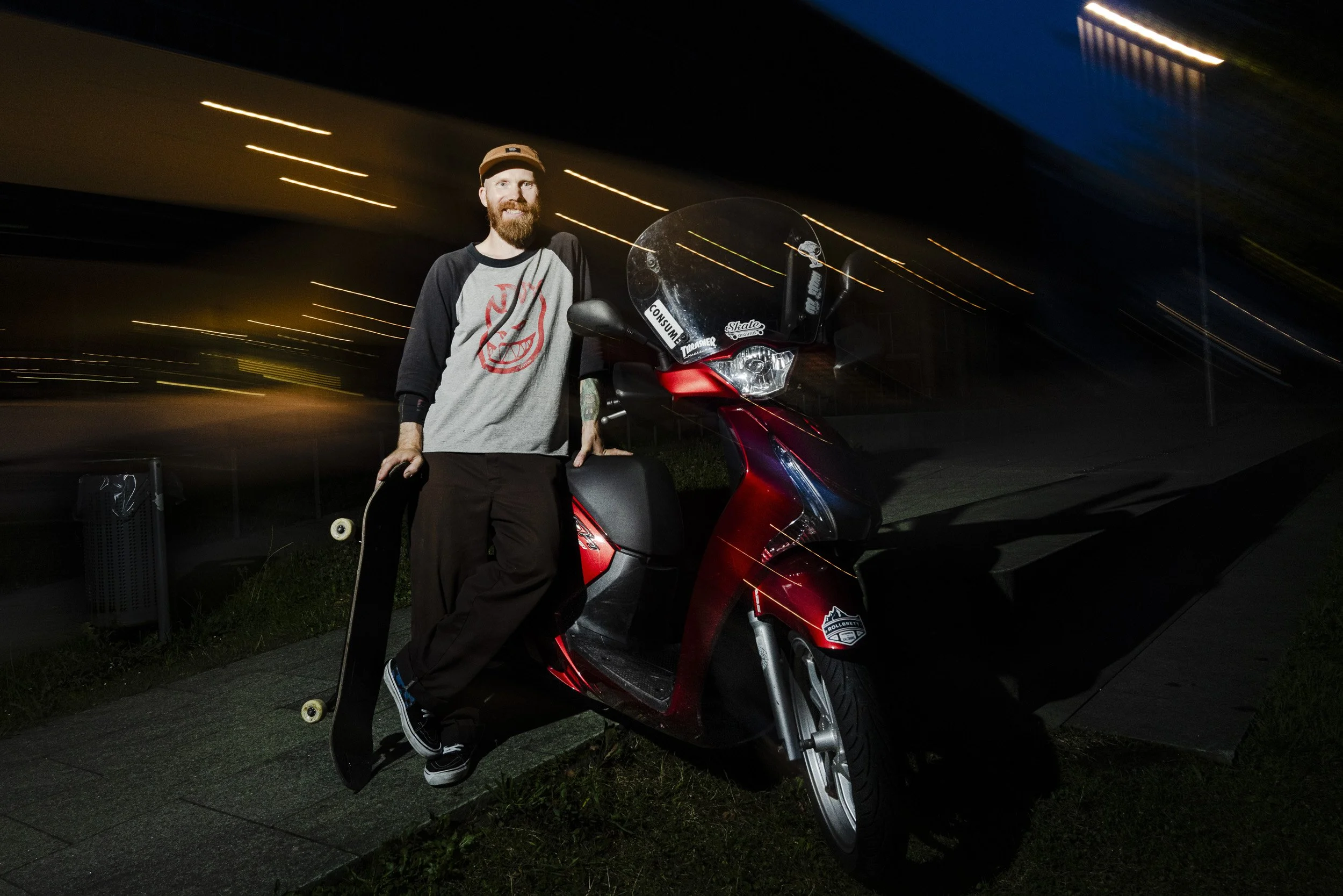 A man with a beard and tattoos, holding a skateboard, leaning on a red scooter at night with blurred street lights in the background.