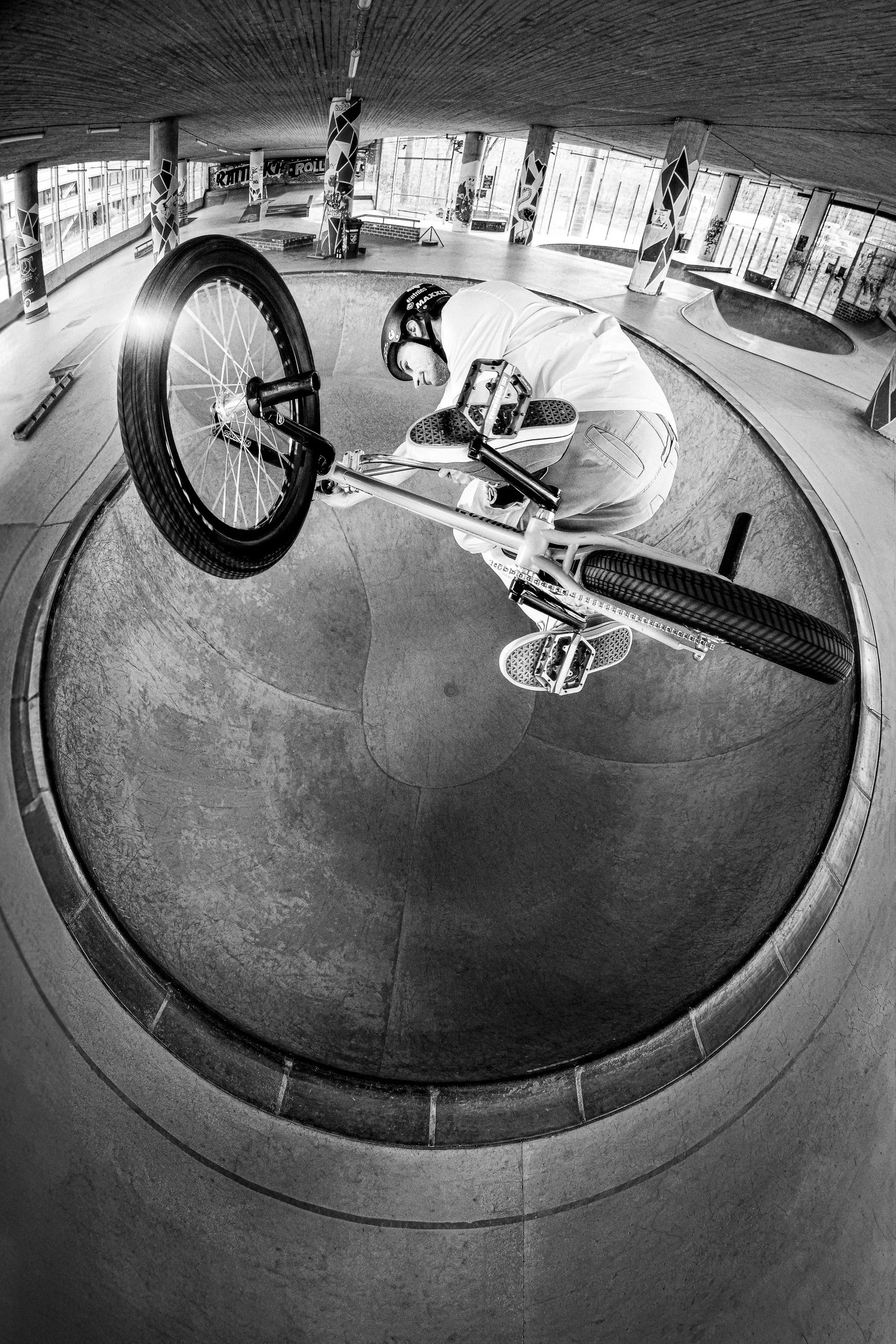 A person in a skatepark riding a BMX bike inside a bowl, wearing a helmet and white clothing, captured with a fisheye lens in black and white.