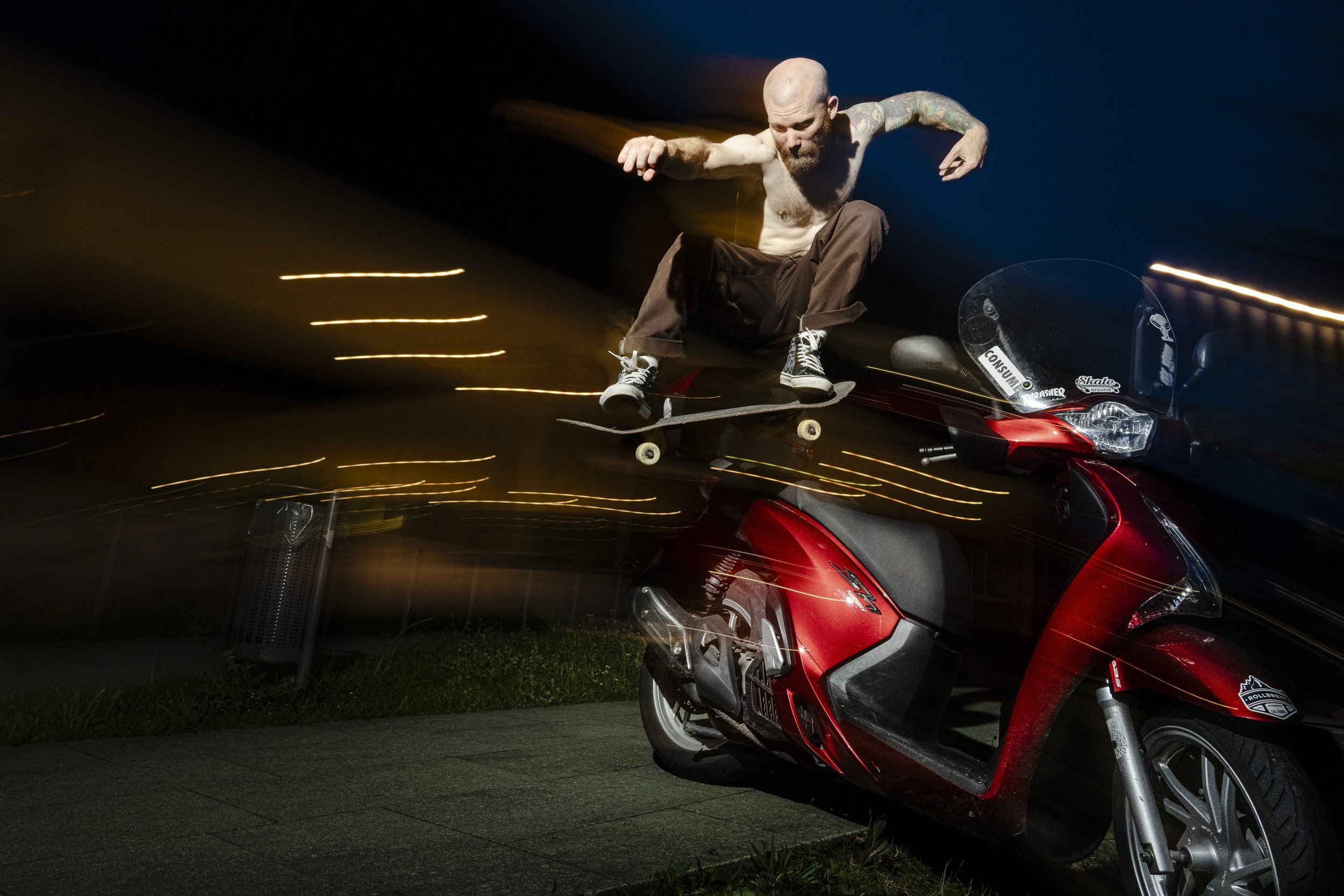 A shirtless man with tattoos skateboarding at night on top of a red scooter.