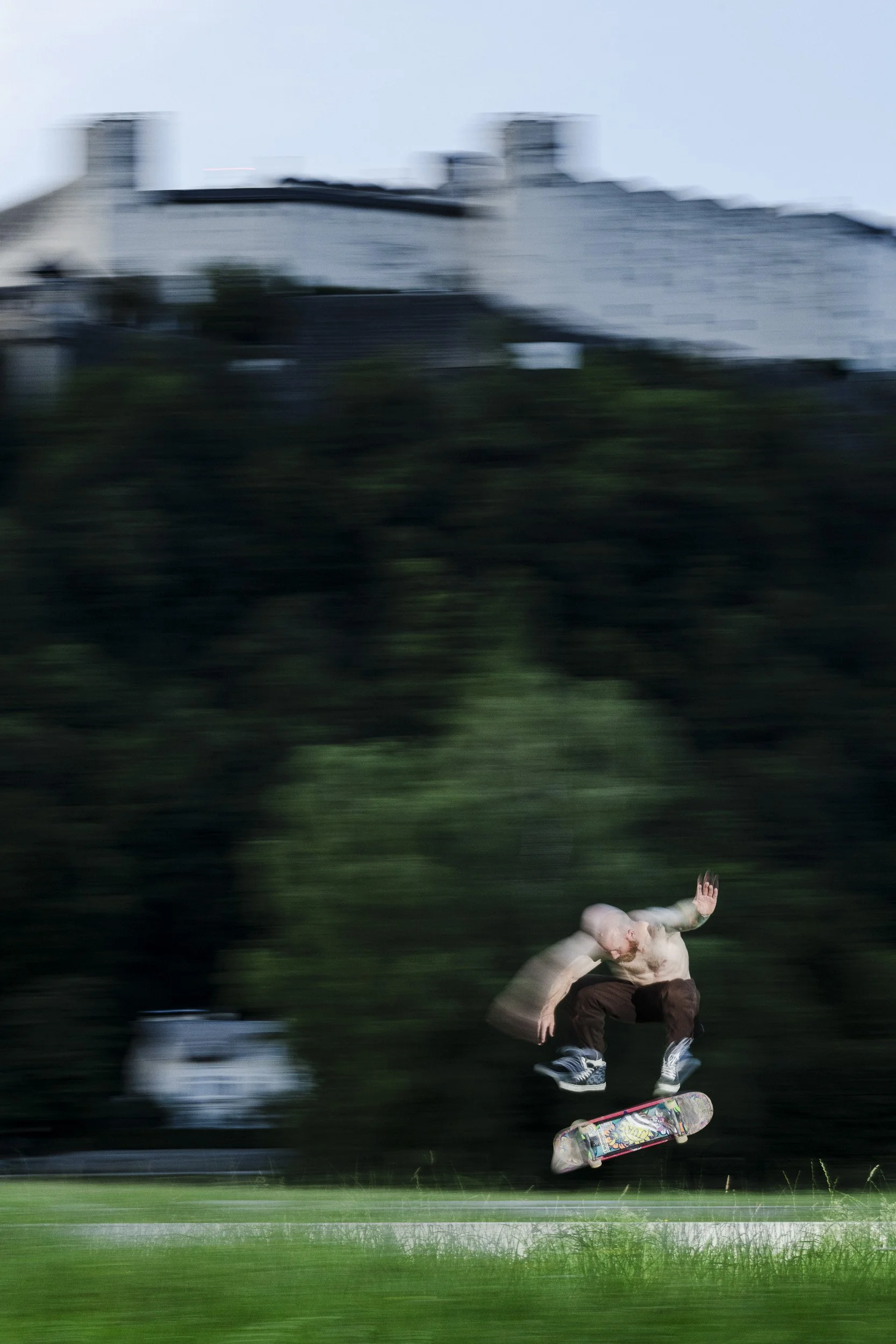 Bald man skateboarding outdoors with motion blur and a house on a hill in the background.