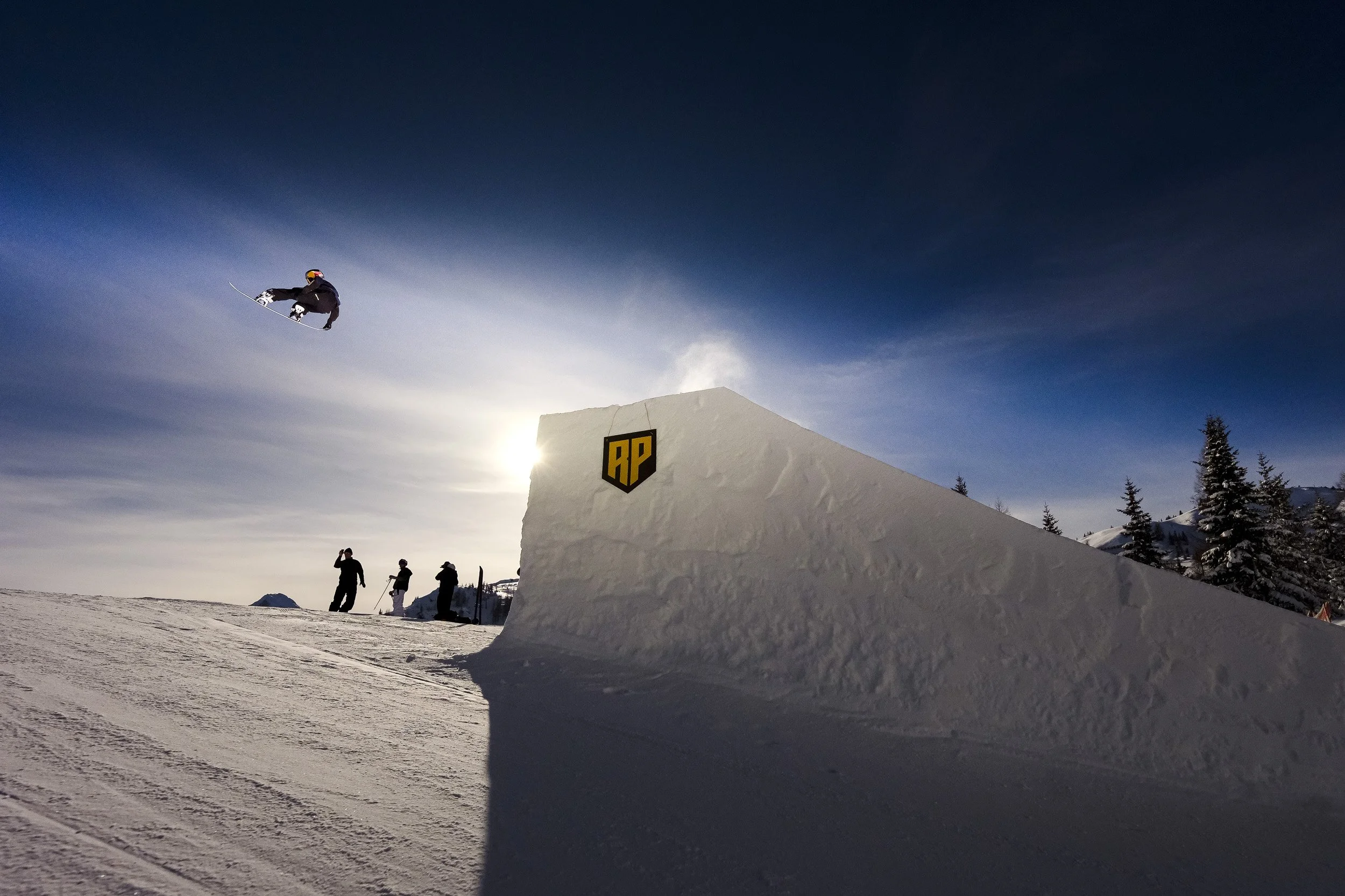 A snowboarder is mid-air performing a jump at a snow park during sunset, with a snow ramp and several onlookers in the background.