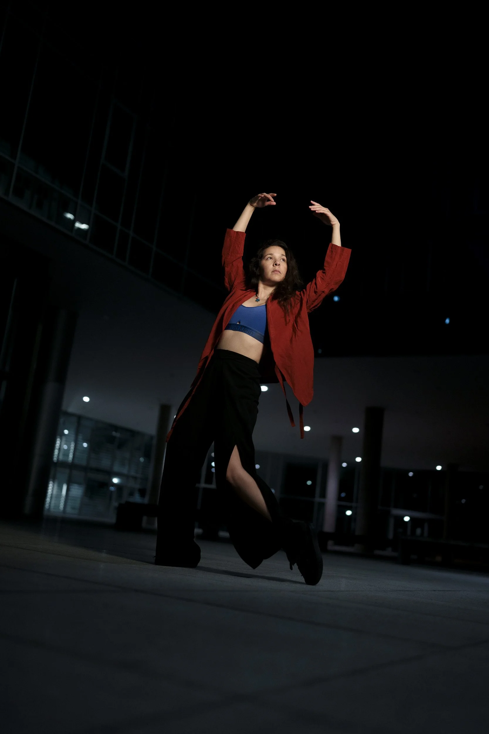 A woman dancing at night outside, wearing a red jacket, blue crop top, and black pants with a slit, in a modern urban setting.