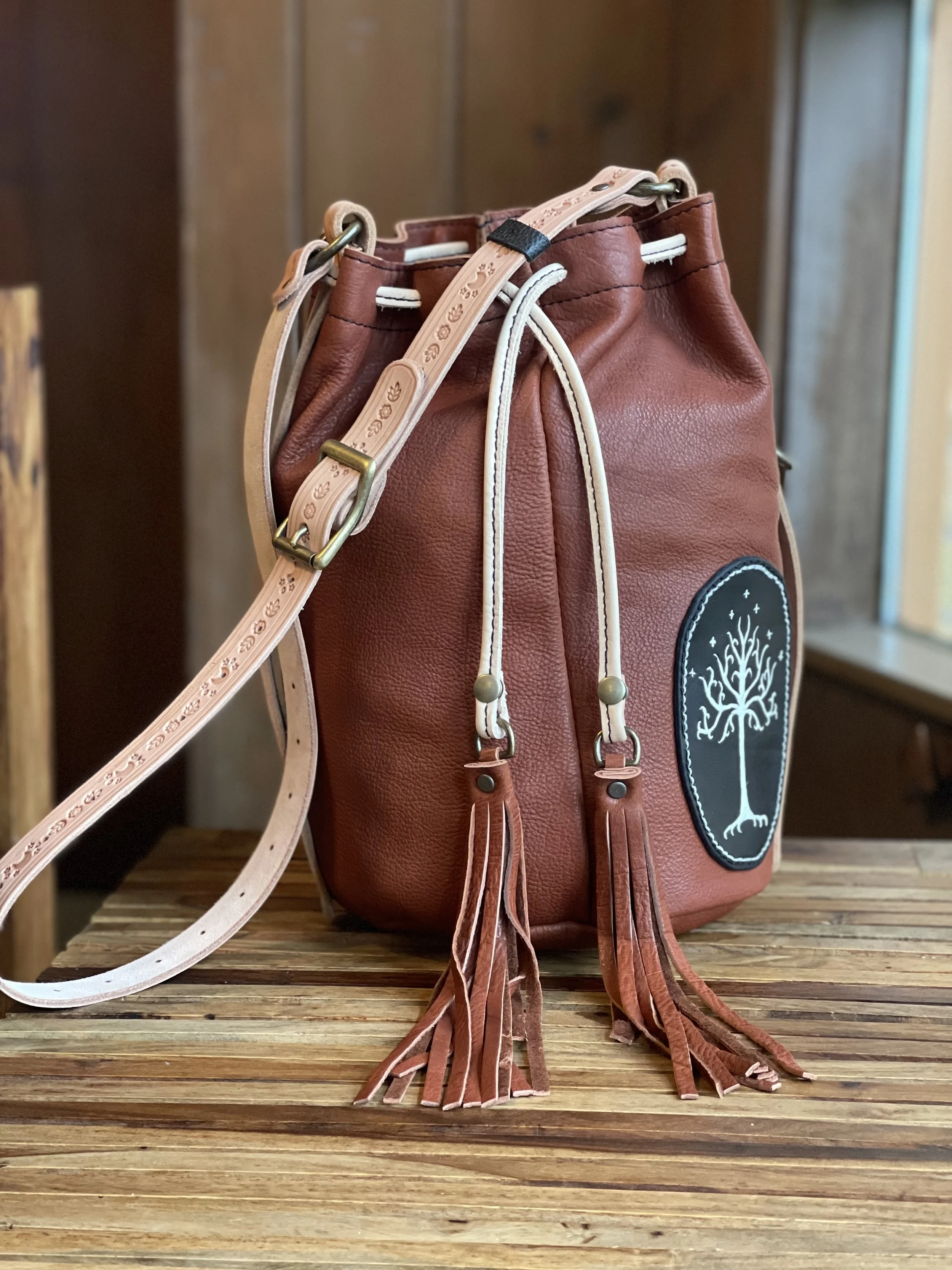 LILI BUCKET BAG IN TOBACCO BISON LEATHER, HAND STAMPED VEGETABLE TANNED LEATHER STRAP, REMOVABLE FRINGE PULLS, AND A CUSTOM REQUESTED SEWN PATCH FROM CUSTOMER'S PERSONAL COLLECTION.