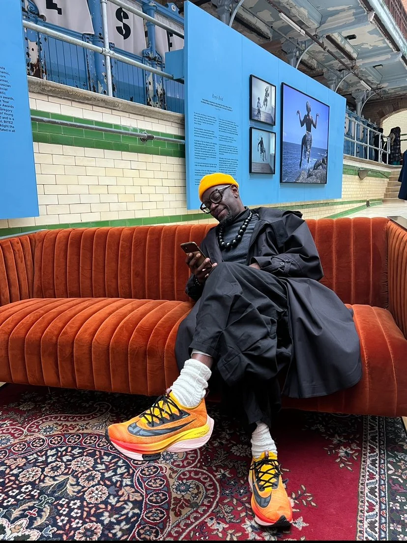 A man wearing a yellow beanie, sunglasses, a black jacket, and black pants sits on an orange velvet sofa in a gallery or museum. He is looking at his phone and is wearing bright orange Nike sneakers with black laces. The background features a blue wall with framed photographs.