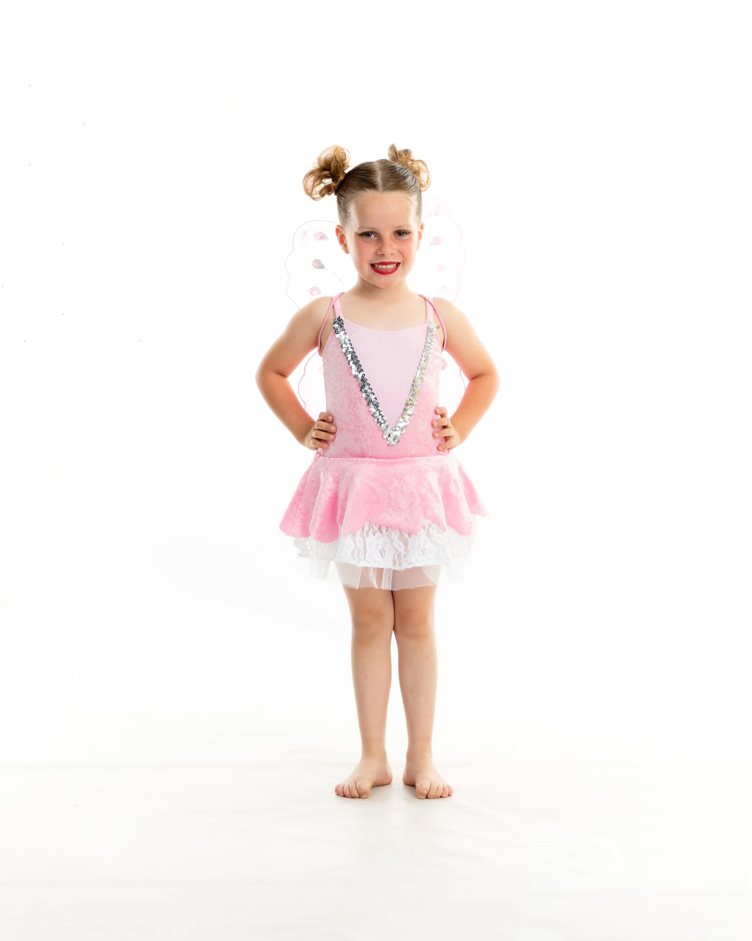 A young girl dressed as a fairy with pink dress and butterfly wings, smiling with hands on hips against a white background.