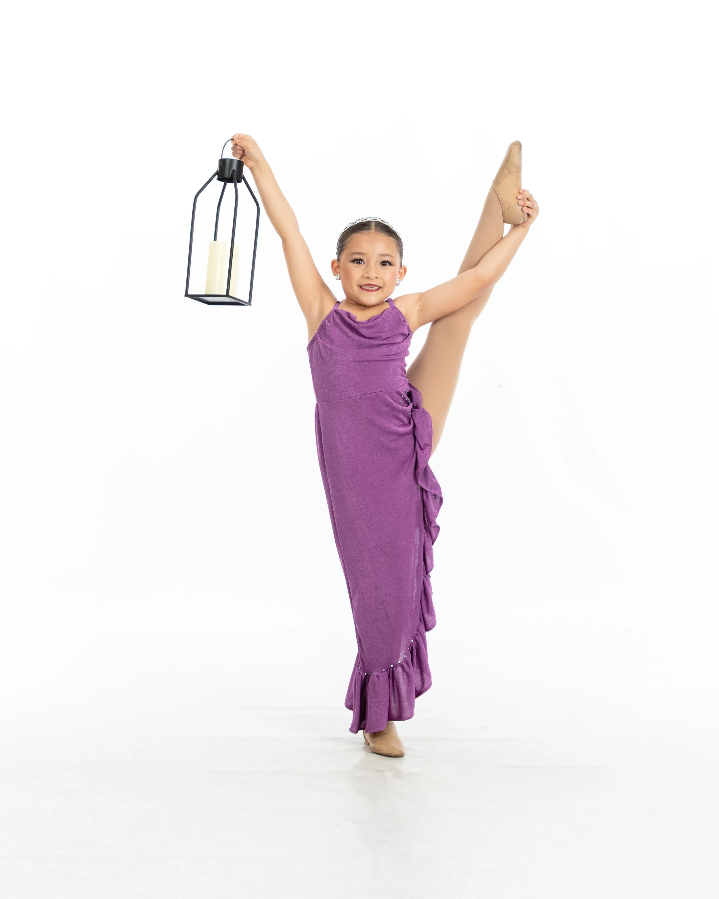 Young girl in a purple dress and beige tights doing a ballet pose, holding a lantern in one hand, standing on one foot against a white background.