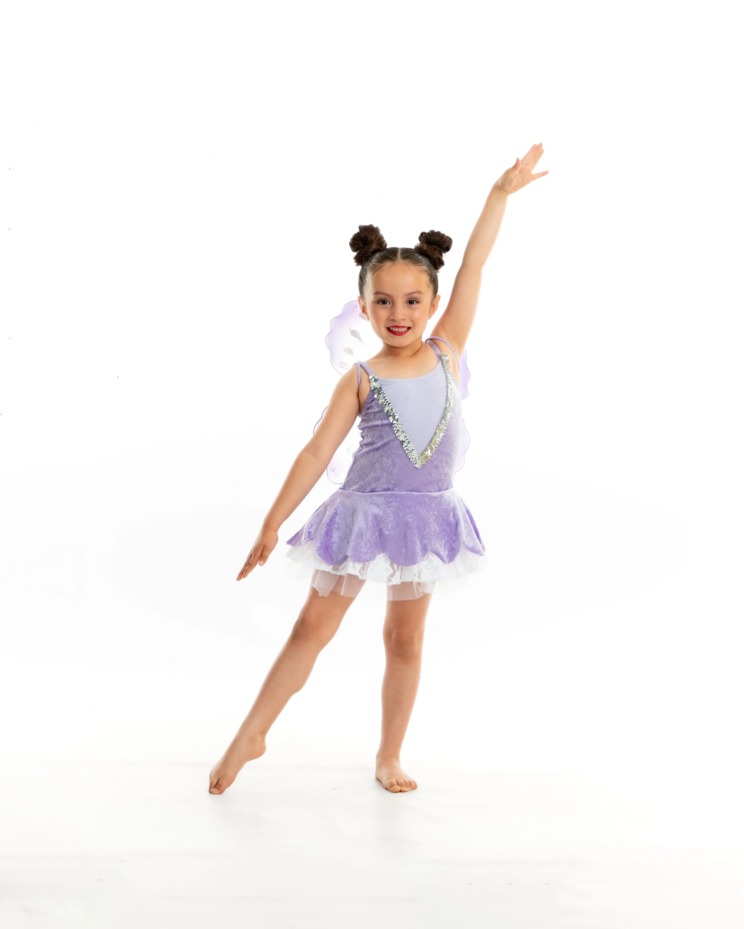 Young girl in a purple fairy costume posing with one arm raised and the other extended sideways, standing barefoot against a white background.