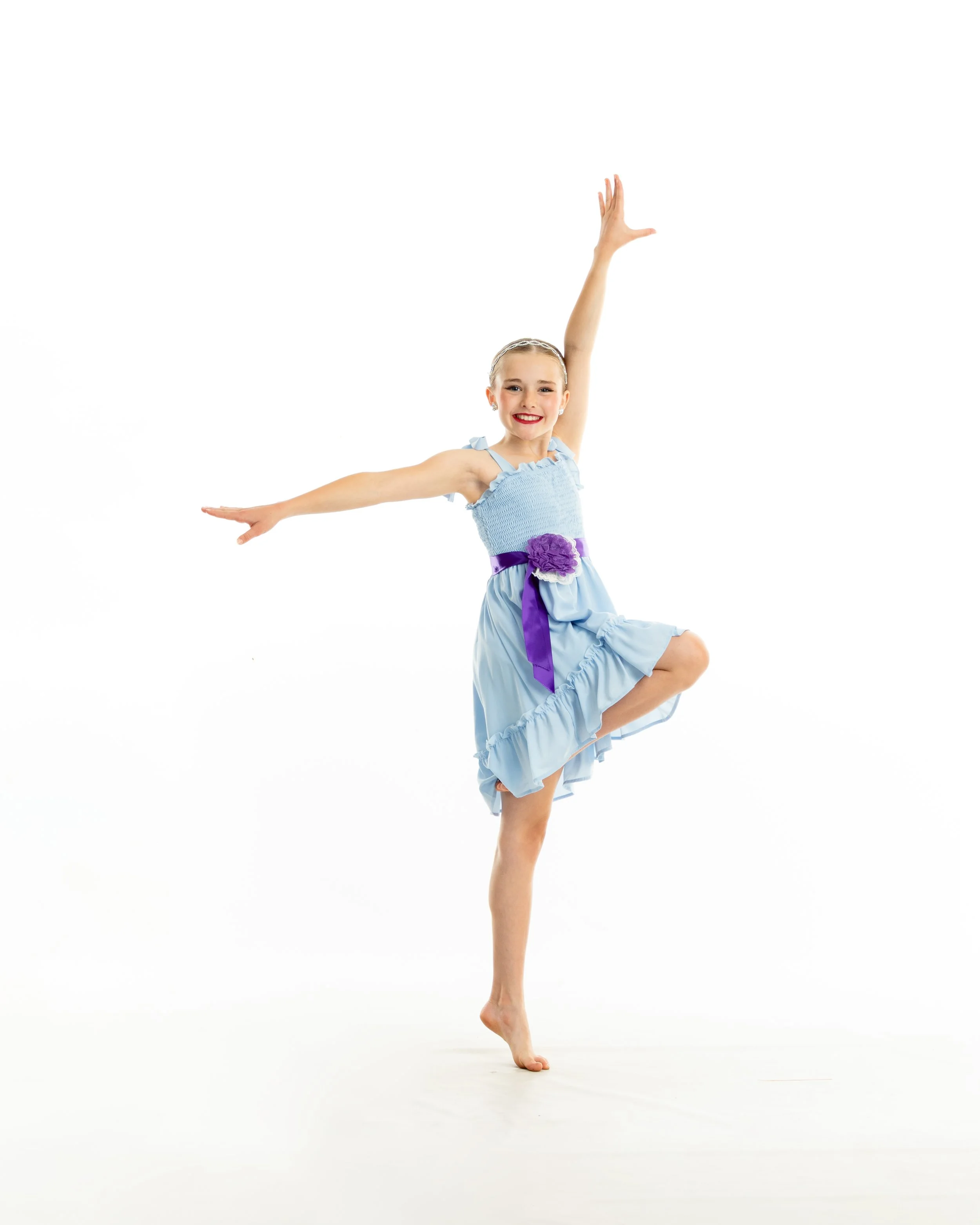 A young girl in a blue dress dancing on one foot with her arms raised, smiling brightly.