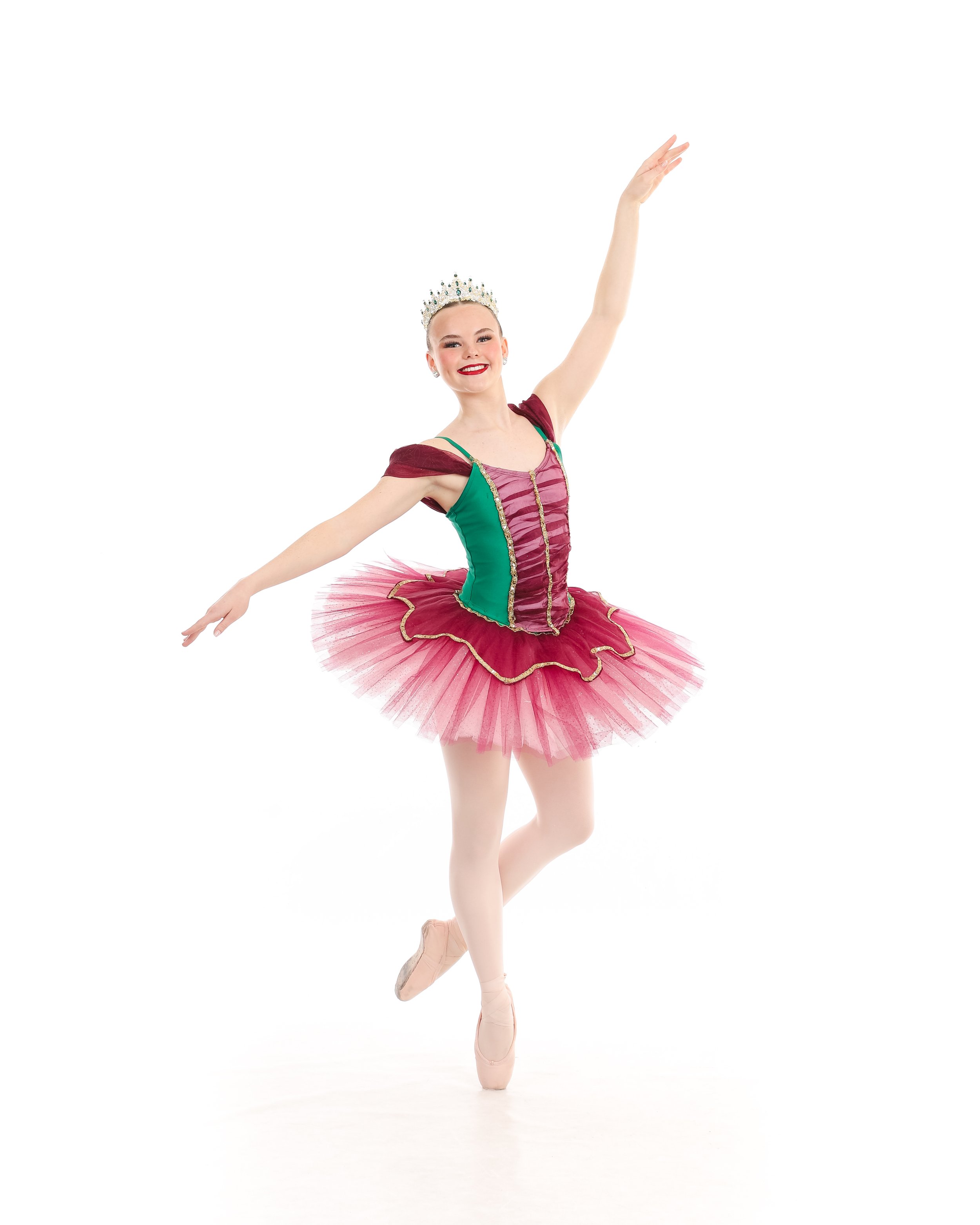 Ballet dancer in a colorful pink and green tutu, wearing a tiara, posing in coupé en pointé shoes against a plain white background.