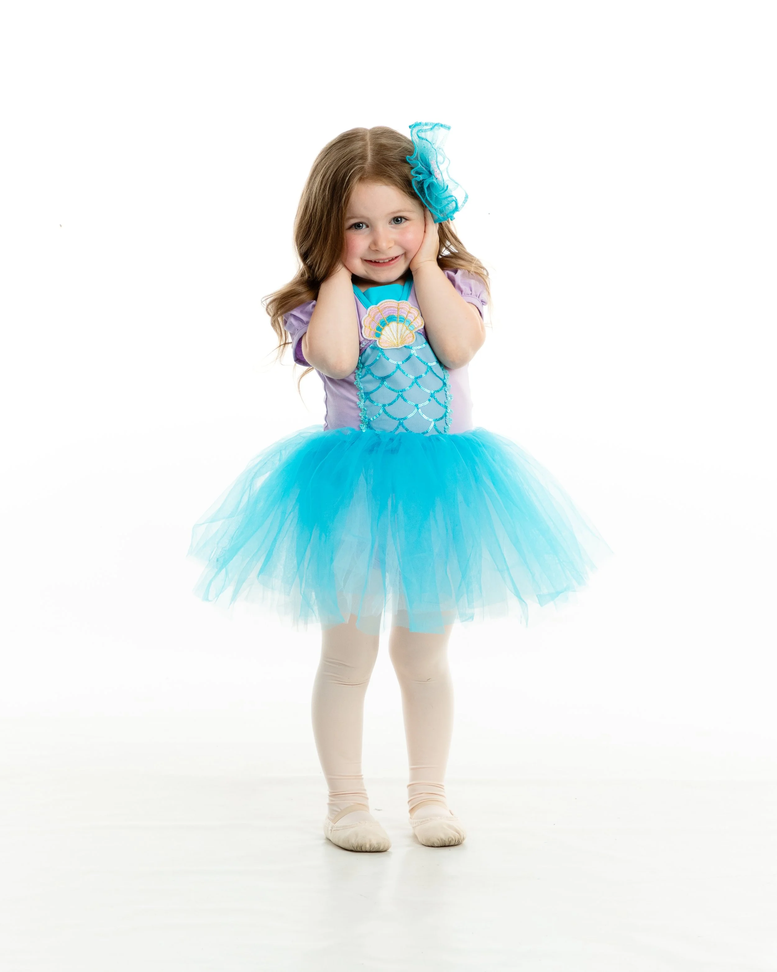 Young girl dressed as a mermaid princess with a purple and blue tutu, teal seashell top, and a blue headband, smiling with hands on her cheeks.