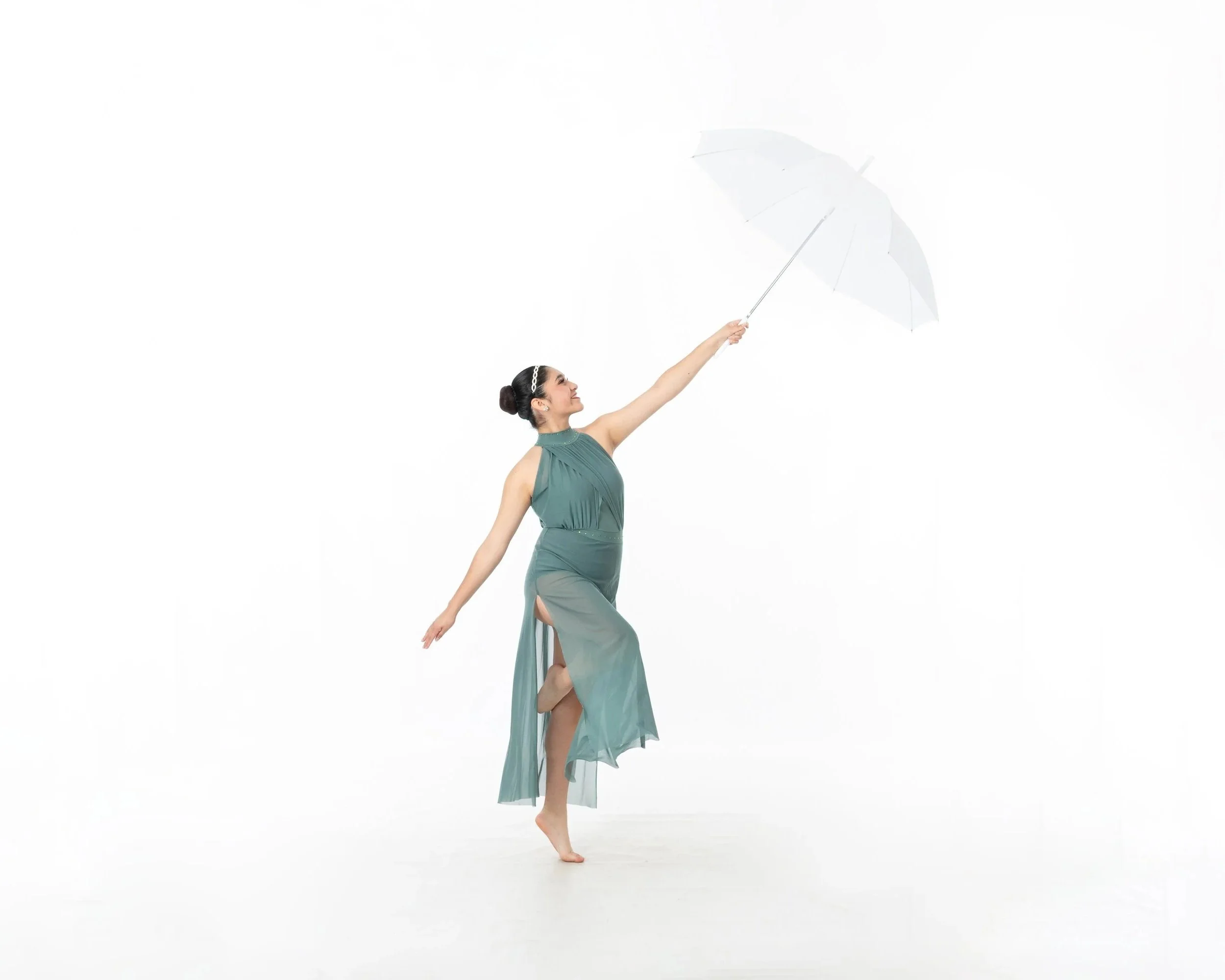 A woman in a teal dress dancing barefoot while holding a white umbrella against a white background.