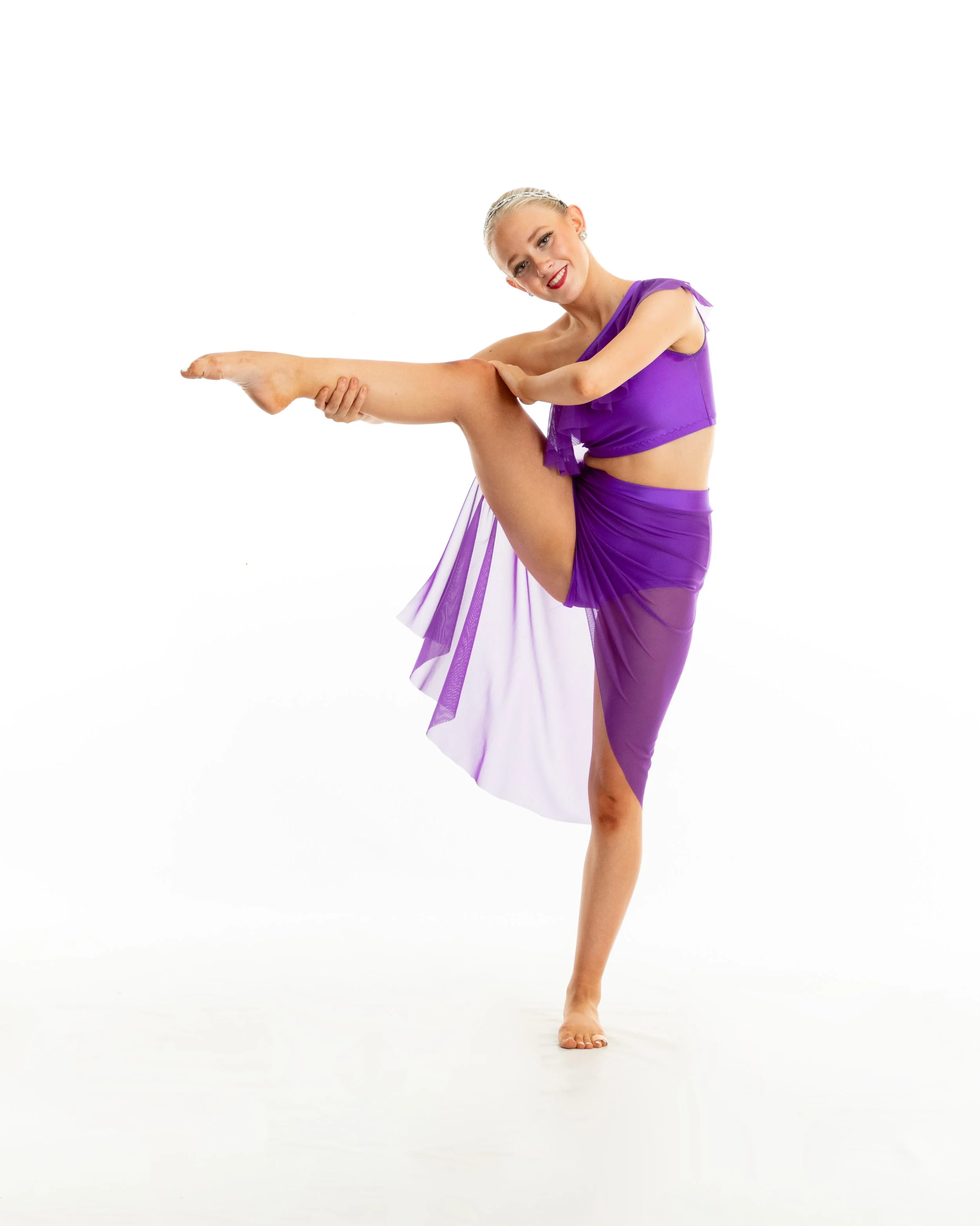 A young woman in a purple dance costume performs a dance pose, balancing on one leg with her other leg raised and extended forward, against a white background.
