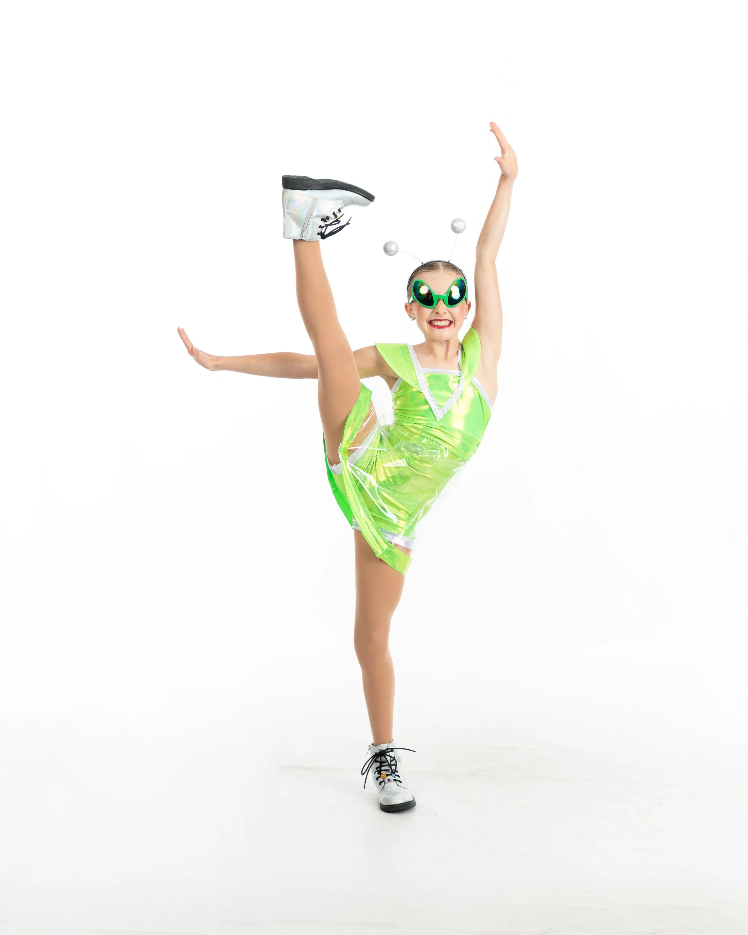 Young girl dressed in a shiny, neon green dance costume, wearing sunglasses with alien faces, performing a high kick in a dance pose against a white background.