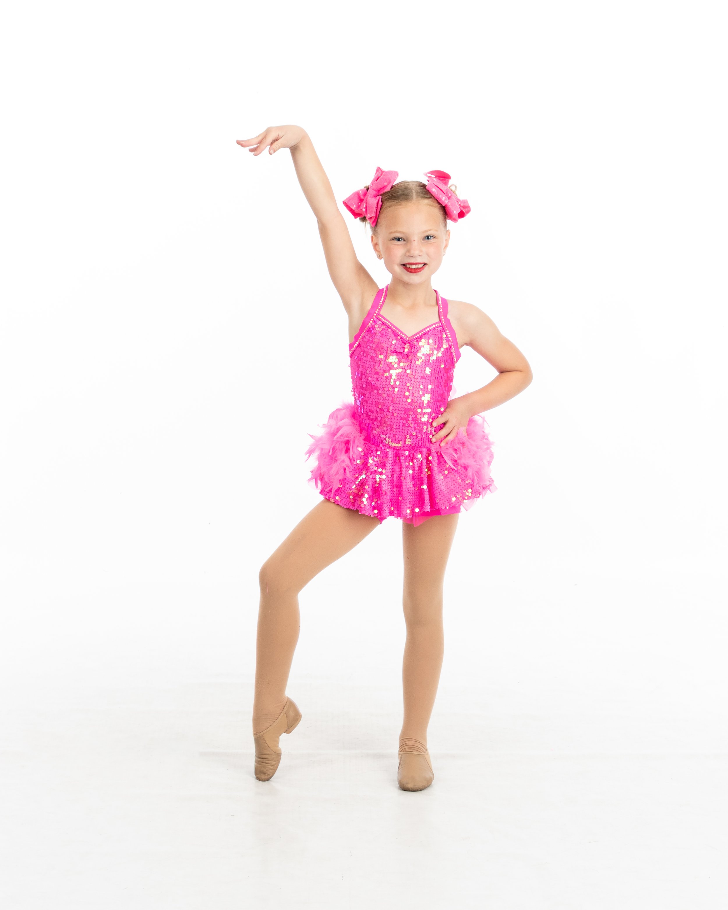 Young girl wearing a bright pink, sequined dance costume with feathered skirt, pink bows in her hair, tan tights, beige dance shoes, posing with one arm raised and smiling.