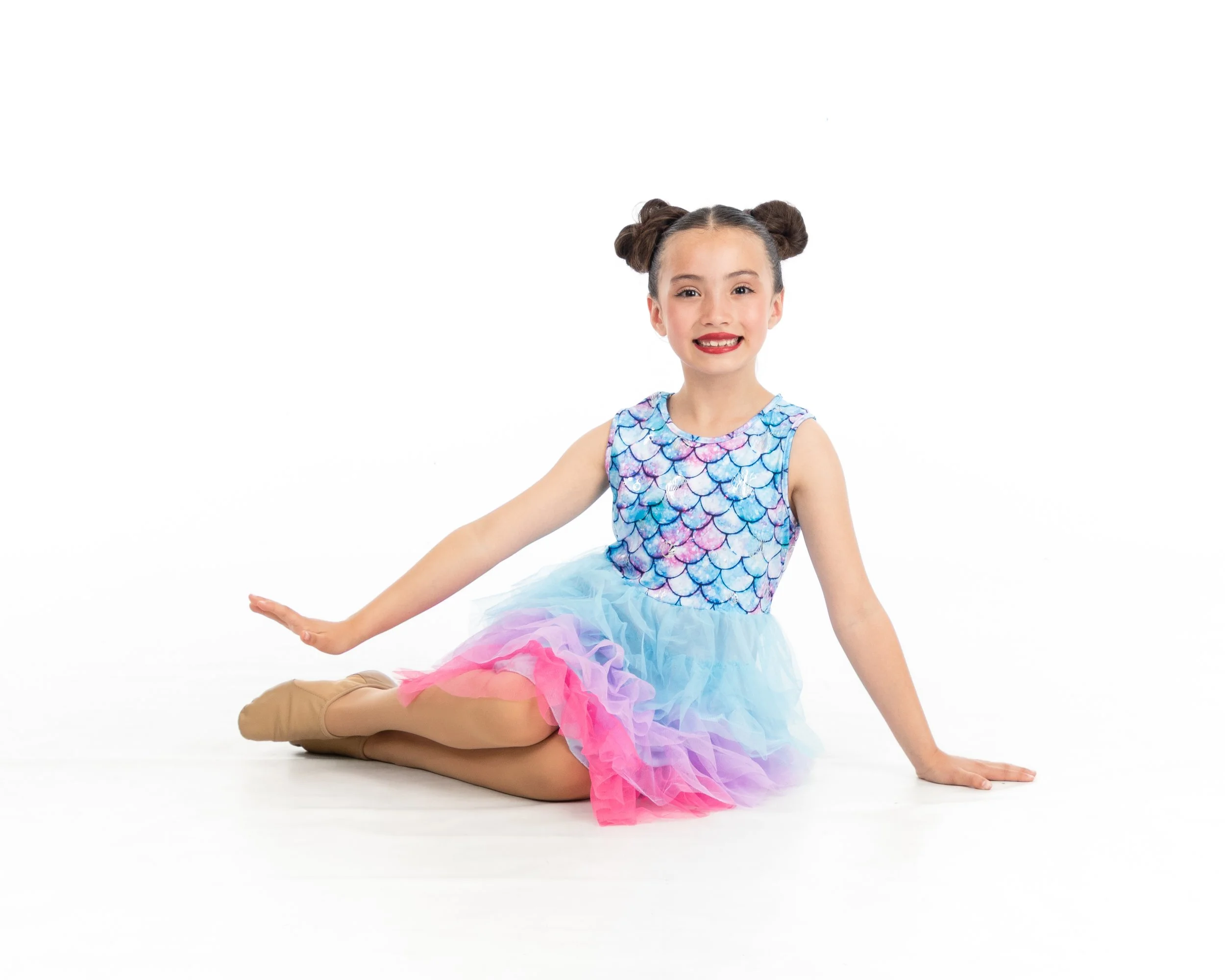 Young girl dressed as a mermaid with a colorful scale top and pastel tutu, posing seated on the floor with one arm extended and smiling.