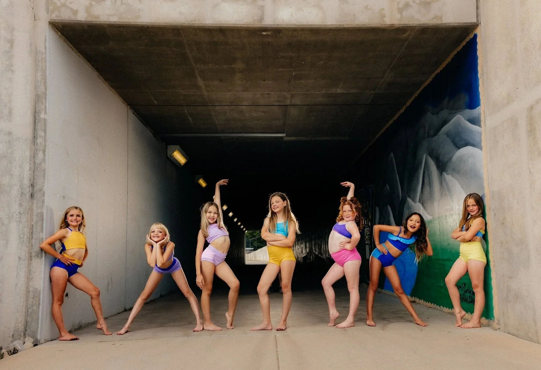 Seven young girls in colorful swimsuits posing under a concrete tunnel with painted murals on the wall.