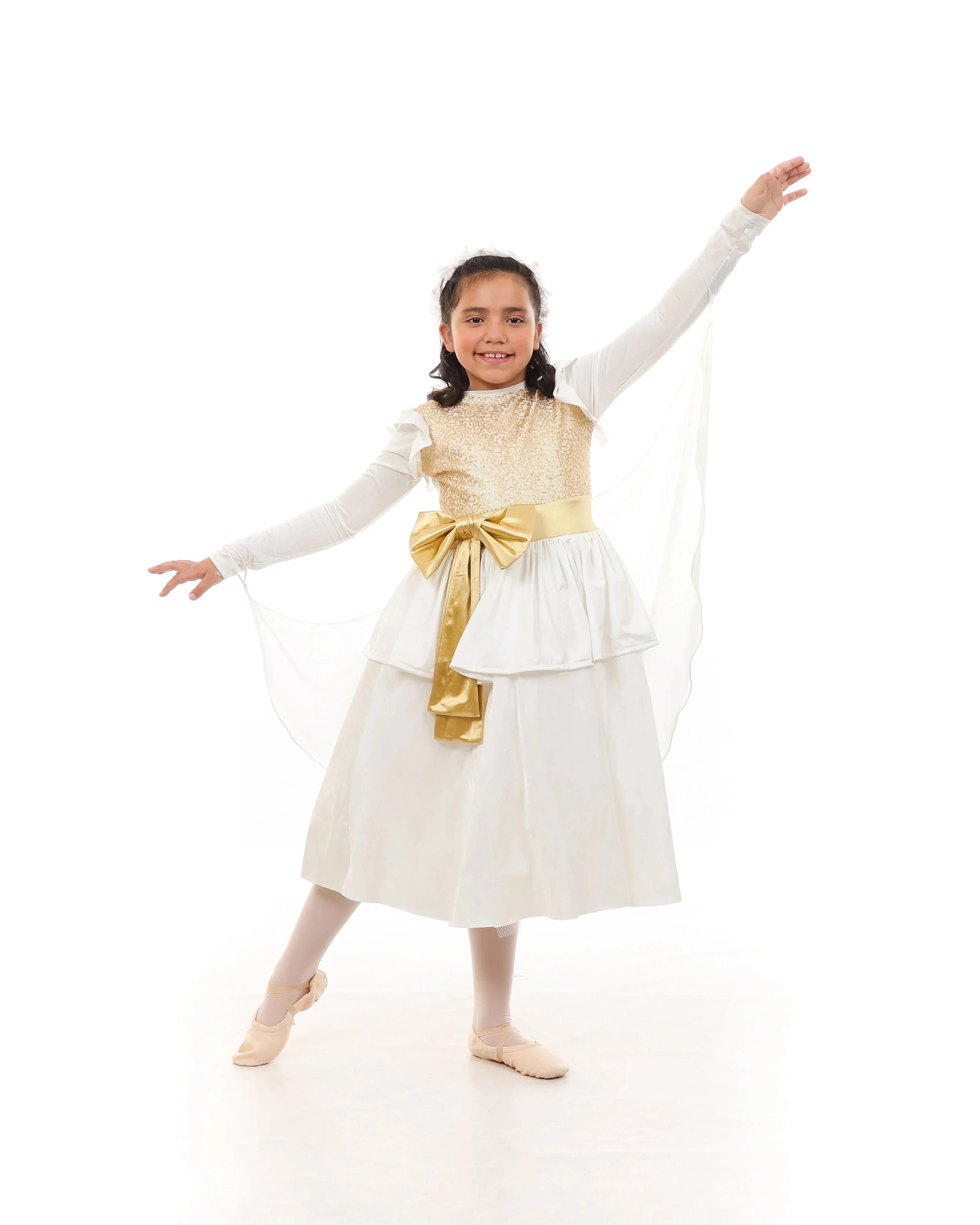 A young girl in a white and gold ballet costume poses on stage, smiling with arms outstretched, against a white background.