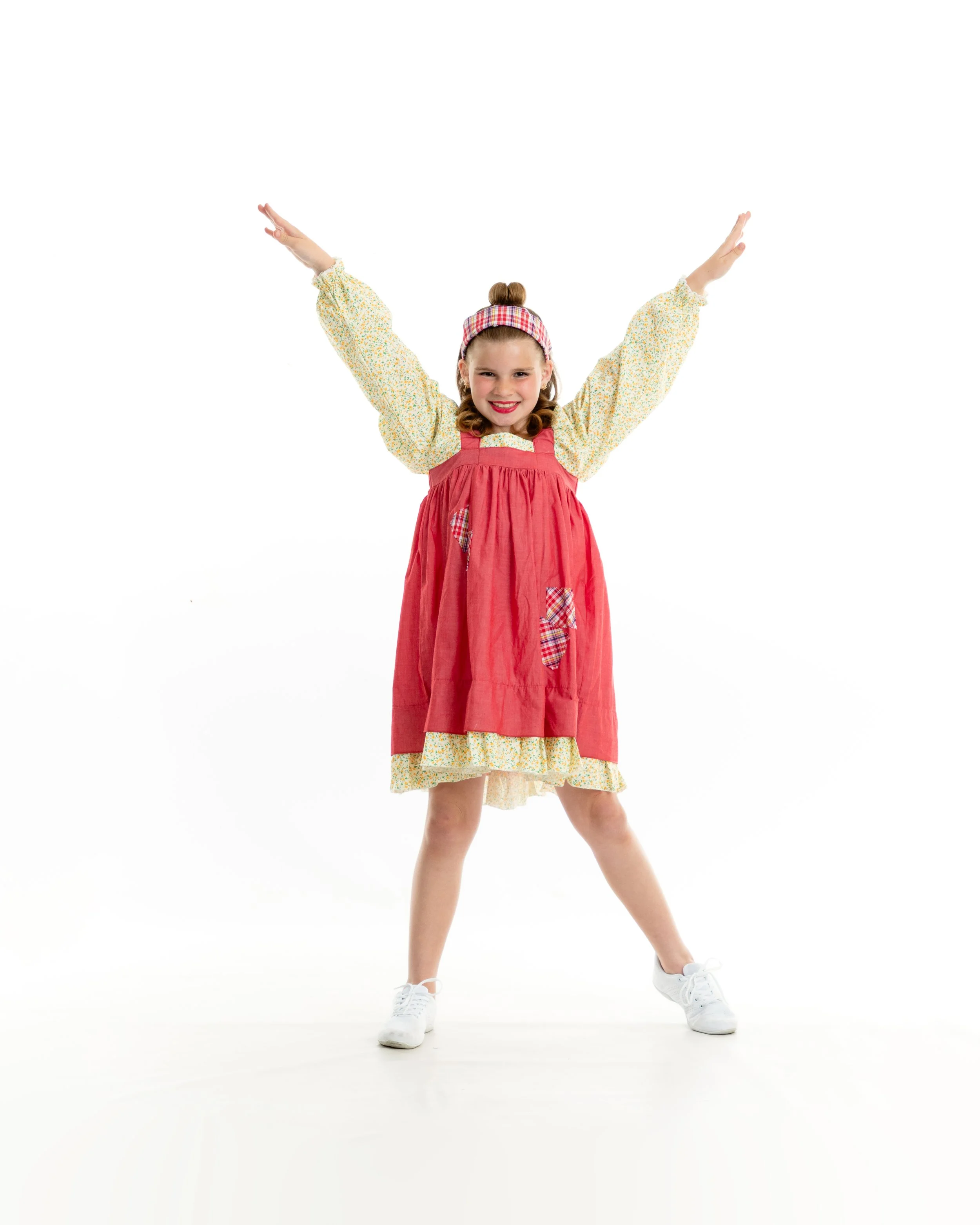 A young girl in a colorful dress and apron, smiling with arms outstretched, standing against a white background.