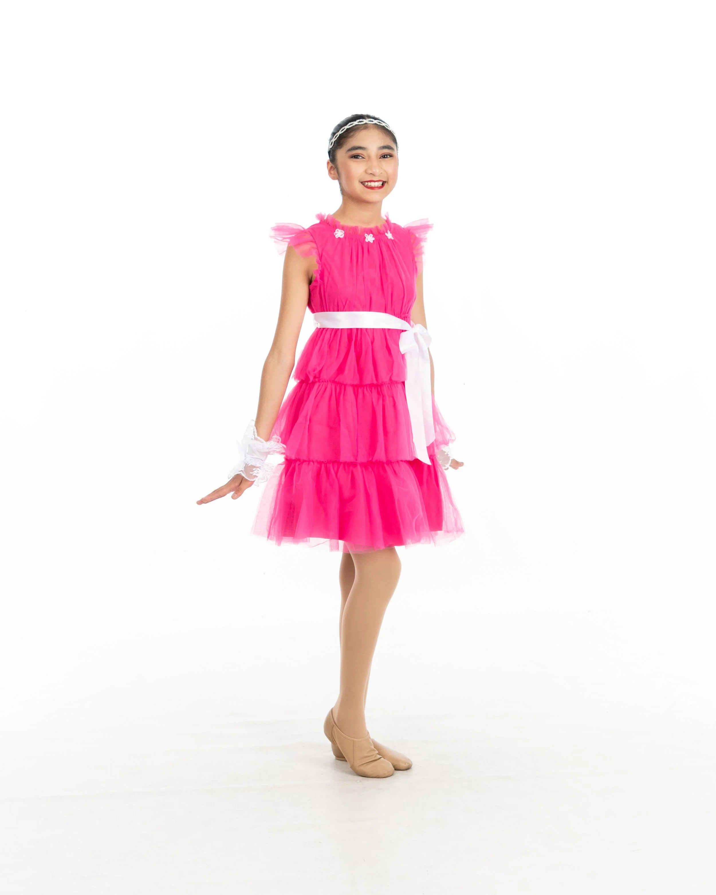 Young girl in a vibrant pink dress with layered tiers, lace cuffs, beige dance shoes, and a white headband, smiling and posing against a plain white background.