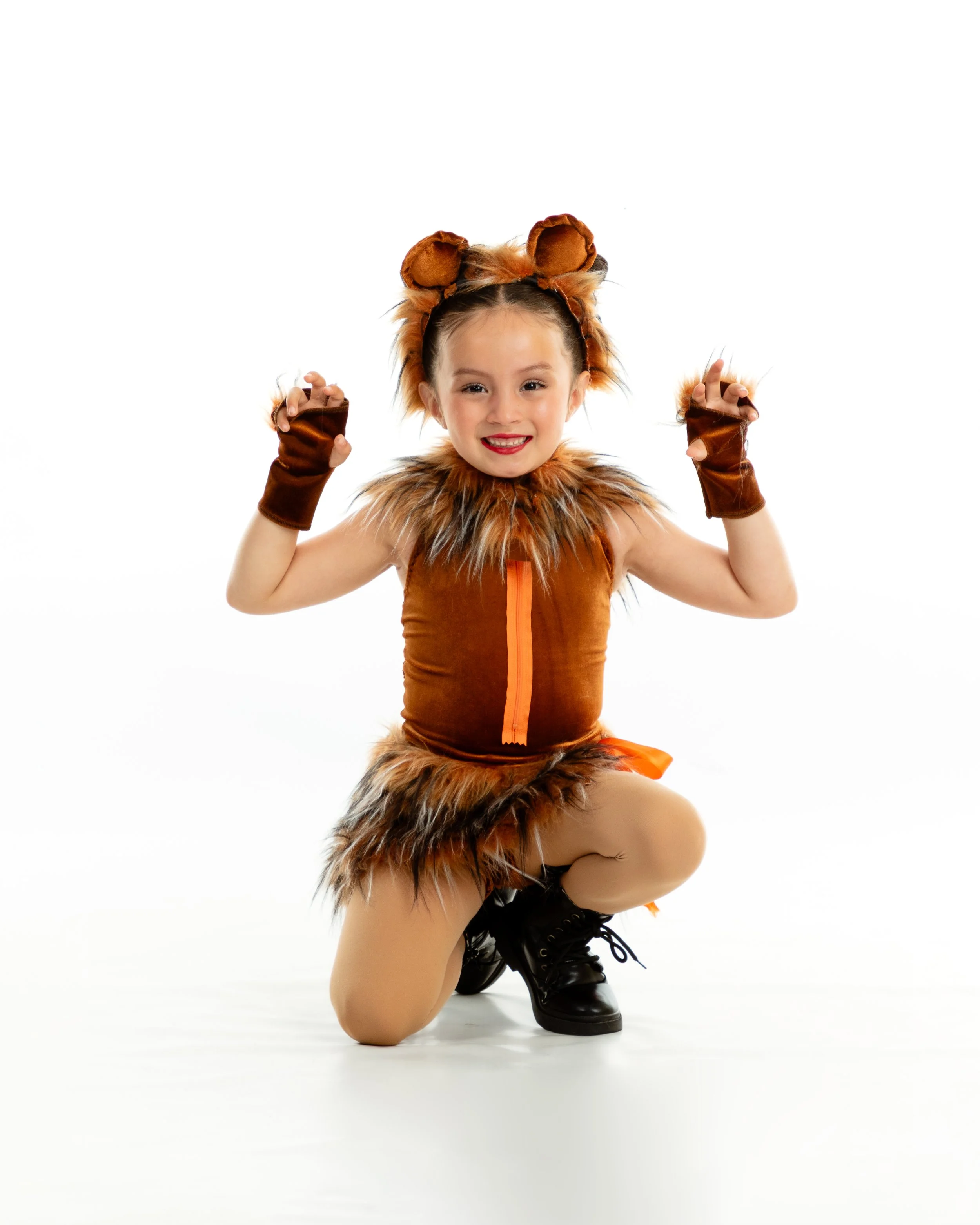 Young girl dressed as a lion, kneeling with arms raised in a posing stance, against a plain white background.