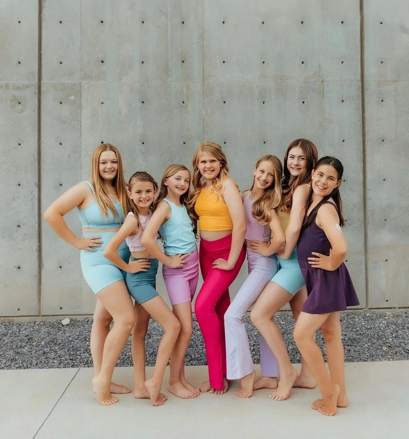 Group of young girls in colorful outfits posing outside against a concrete wall.