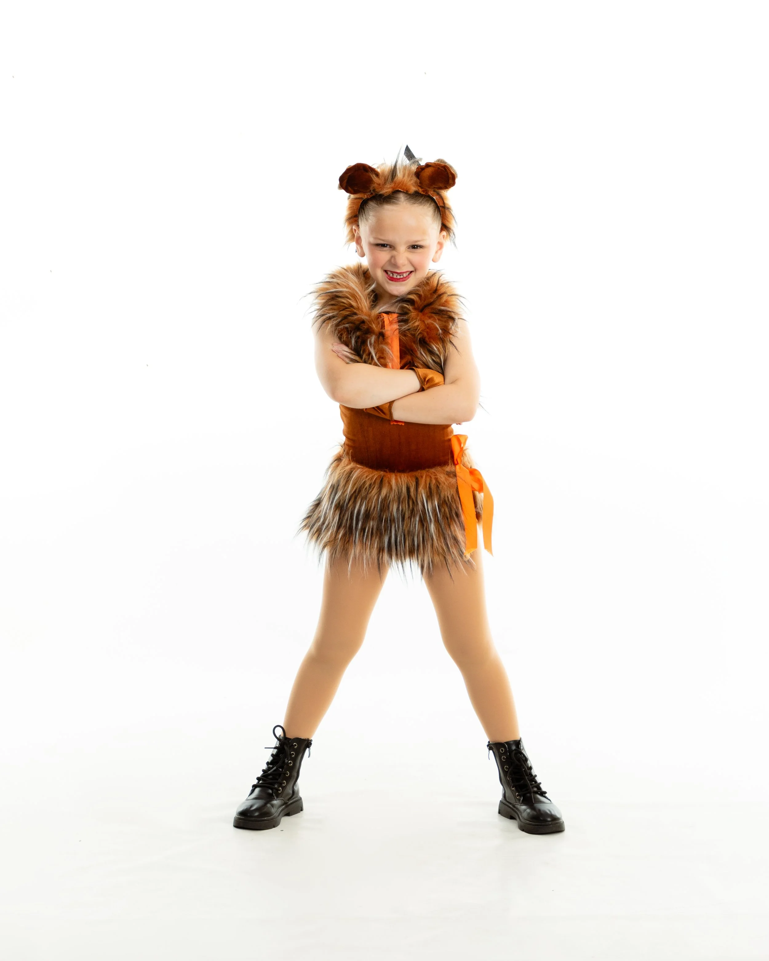 Young girl in a lion costume with a furry mane, arms crossed, standing with legs apart, smiling, against a white background.