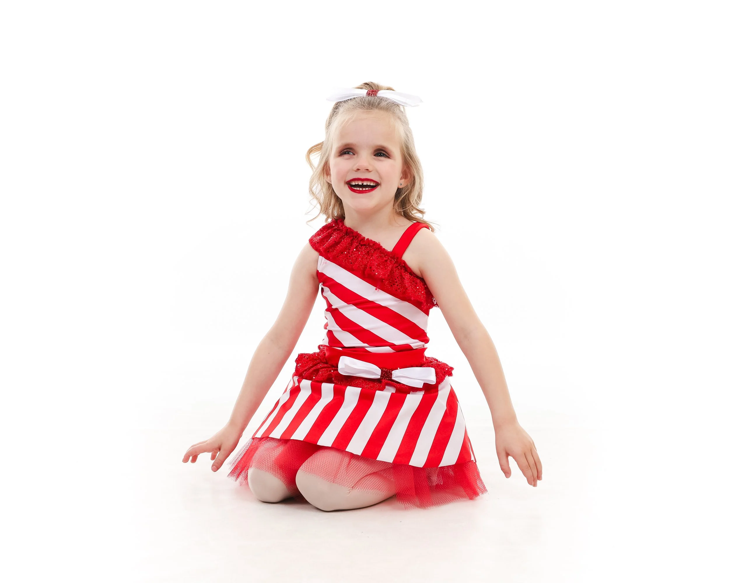 Smiling young girl in red and white striped dress with tulle skirt, kneeling on the floor, white background.