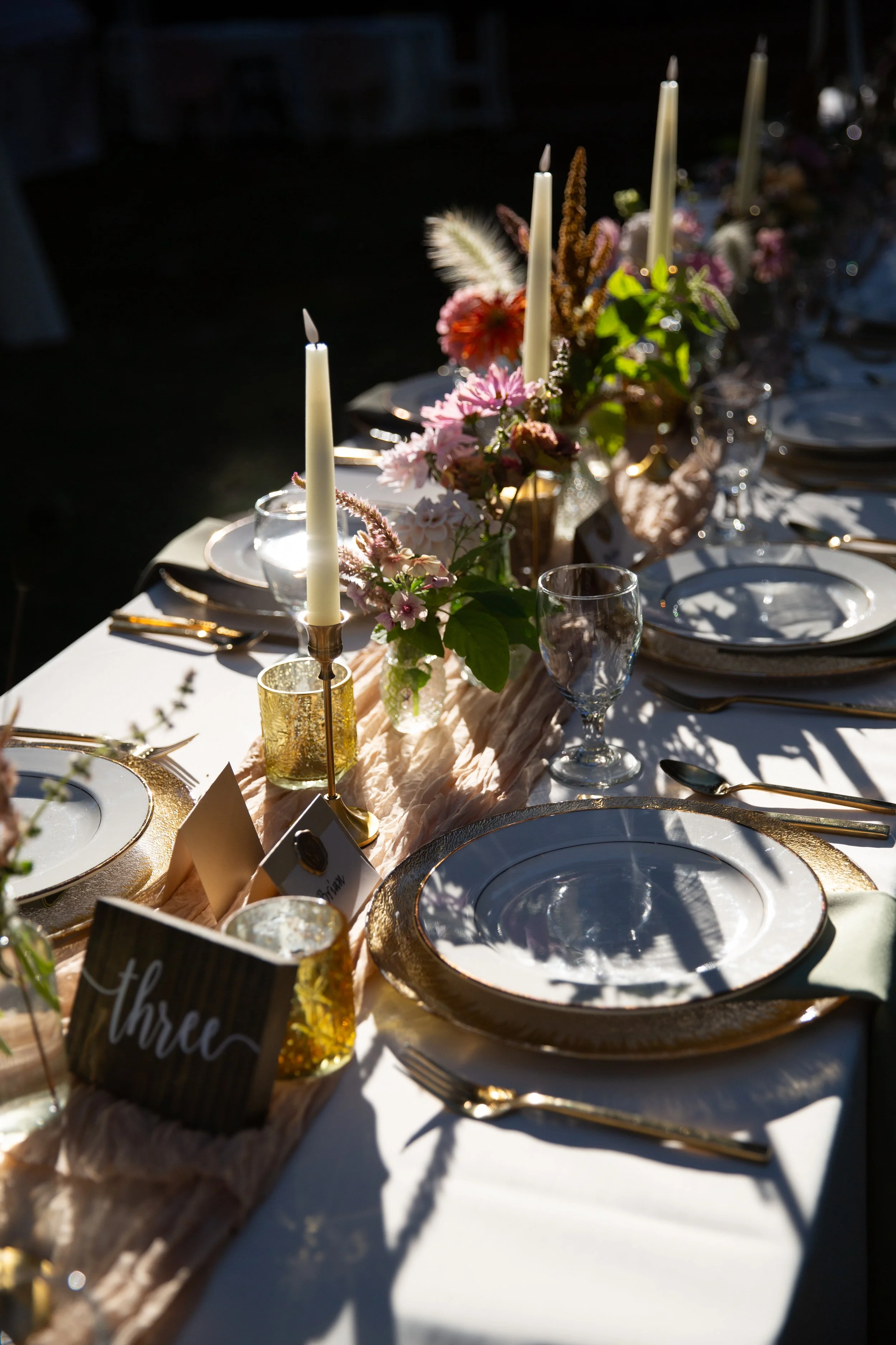 A decorated outdoor dinner table during a wedding reception in Lisbon with tall white candles, pink flowers, green leaves, glassware, and gold-rimmed plates, set in natural sunlight.