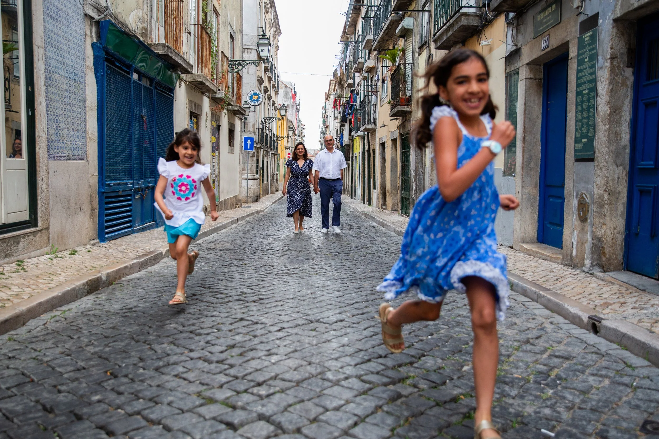 Playful and natural family photoshoot in Lisbon with two kids running down an historic street in Principe Real while their parents smile watching them from the background.