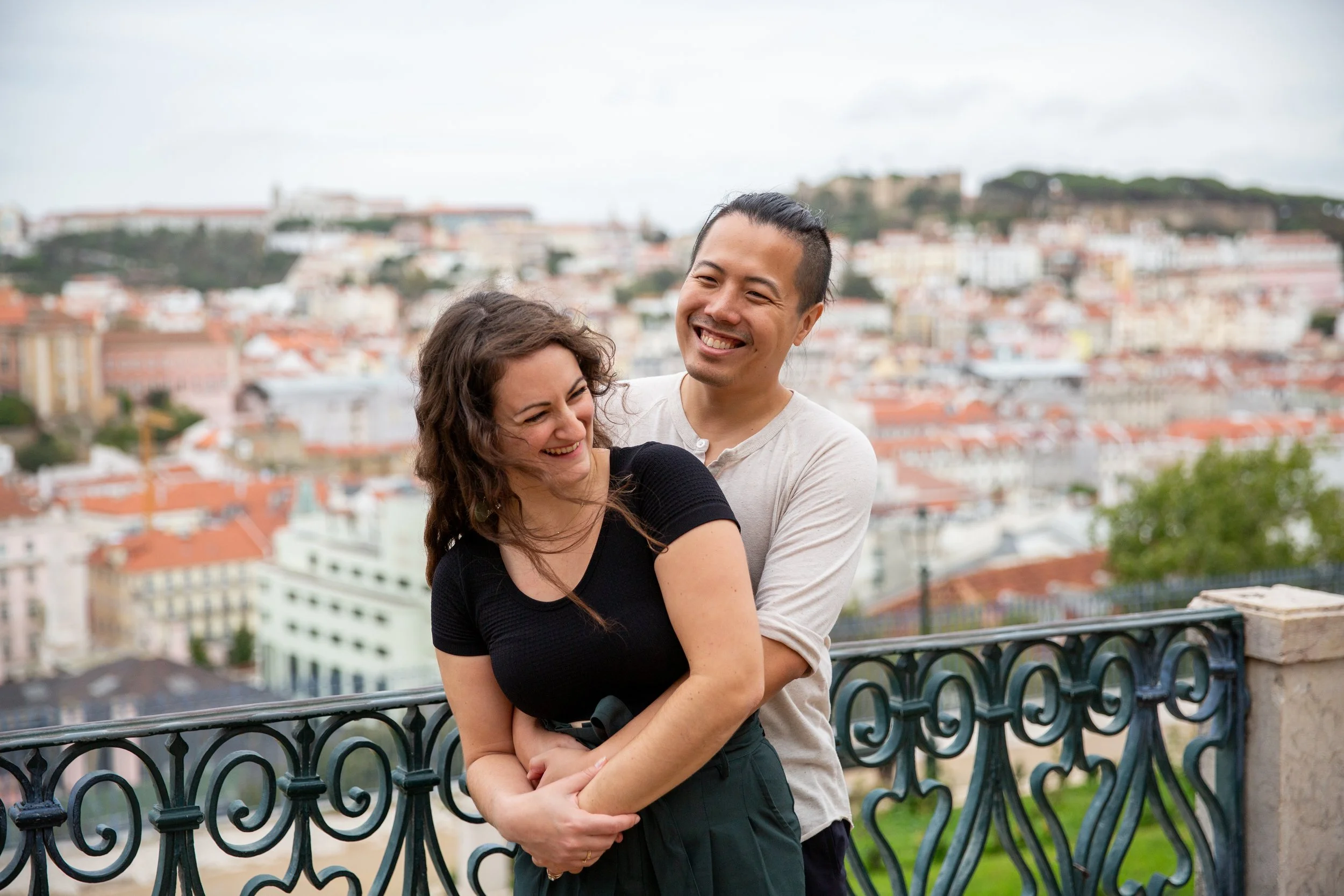 A smiling couple hugging on at the viewpoint looking over the city of Lisbon at the Miradouro de São Pedro de Alcântara.