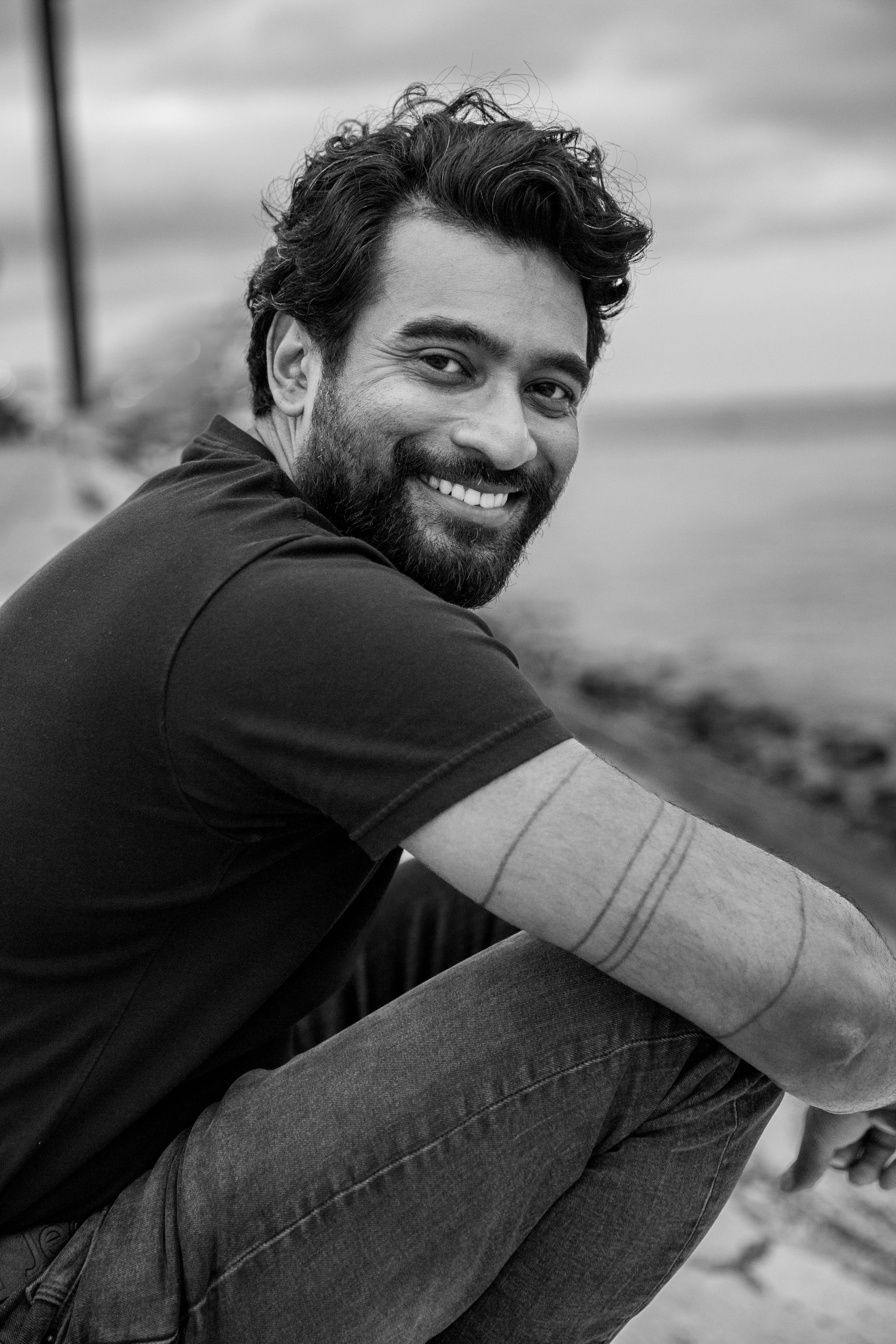 Black and white portrait of a man with a beard, smiling, sitting outdoors by the Tagus River in Lisbon during a personal branding photoshoot in Lisbon.